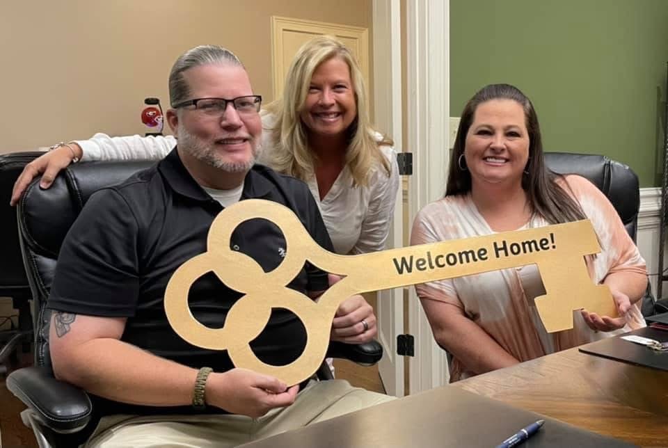 Three smiling people sitting at a table, one holding a large cardboard key with a sign that says 'Welcome Home!' on it, in an indoor setting.