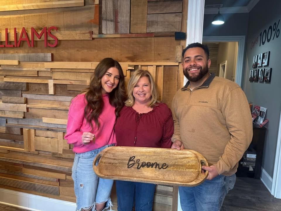 Three people standing indoors, holding a wooden tray with 'Broome' written on it, smiling at the camera, in front of a wooden wall with the partial word 'WILLIAMS' visible.