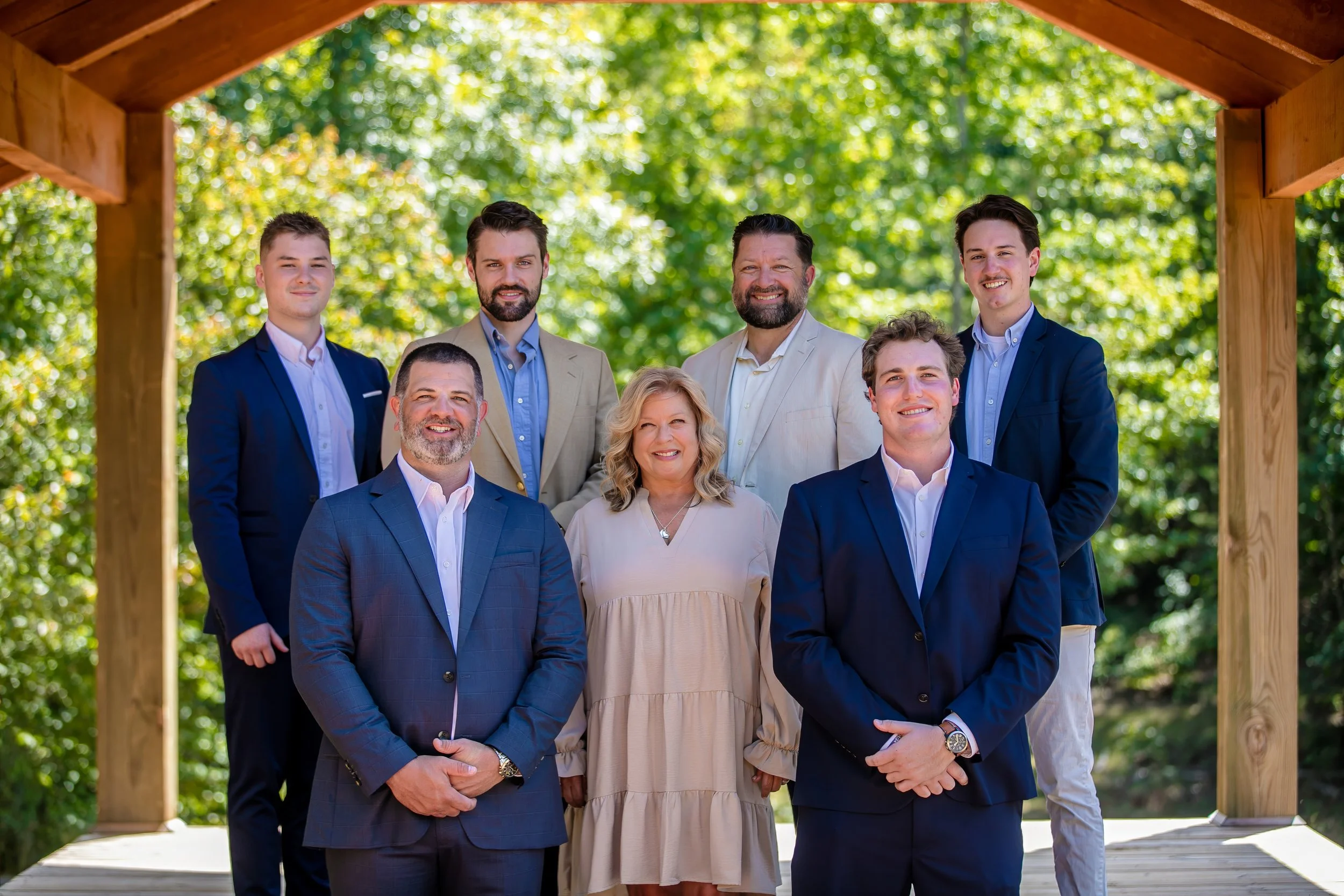 Group of eight people, six men and two women, standing outdoors on a wooden platform under a gazebo with green trees in the background. All are dressed in business casual or formal attire, smiling for the photo.