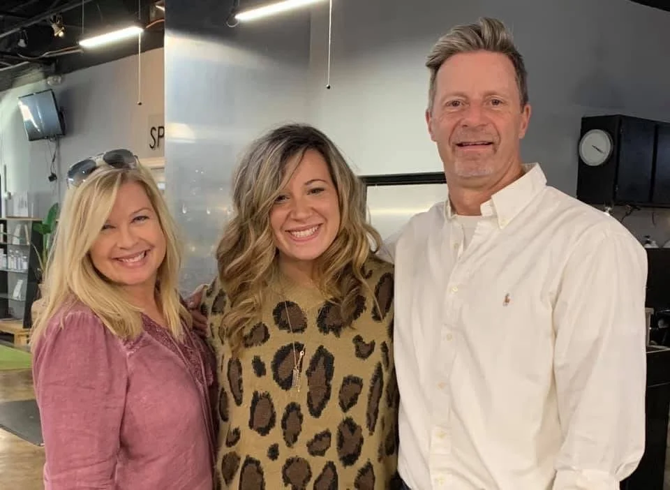 Three smiling adults standing together in an indoor space, with a woman on the left wearing a pink top, a woman in the middle wearing a leopard print top, and a man on the right wearing a white shirt.