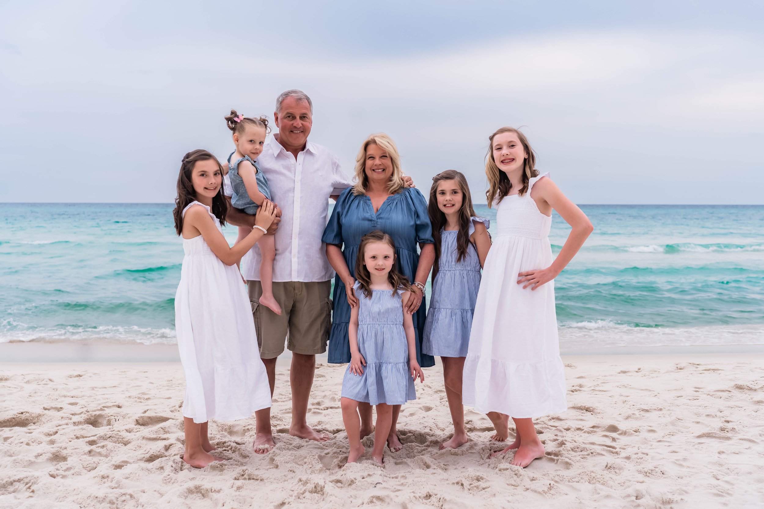 Family of seven at the beach, standing on sand with ocean in the background, smiling and posing for the photo.