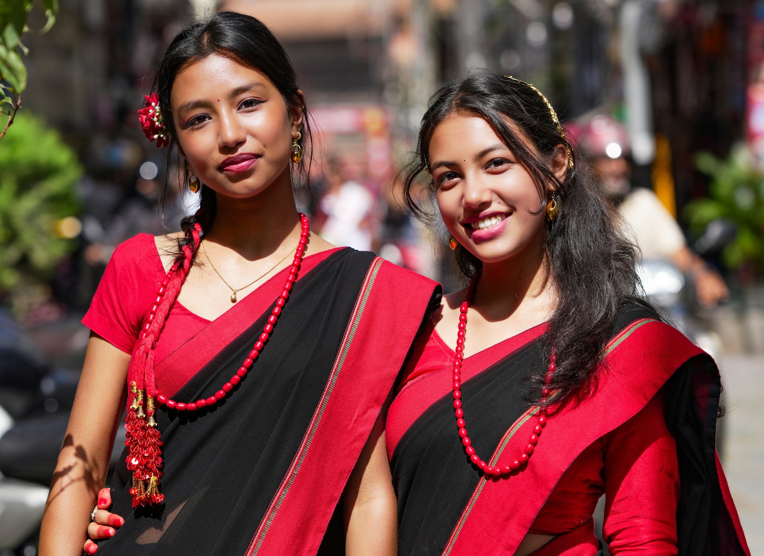 Two young women dressed in traditional Indian sarees with red and black colors, wearing red beaded necklaces and earrings, standing outdoors on a sunny day.