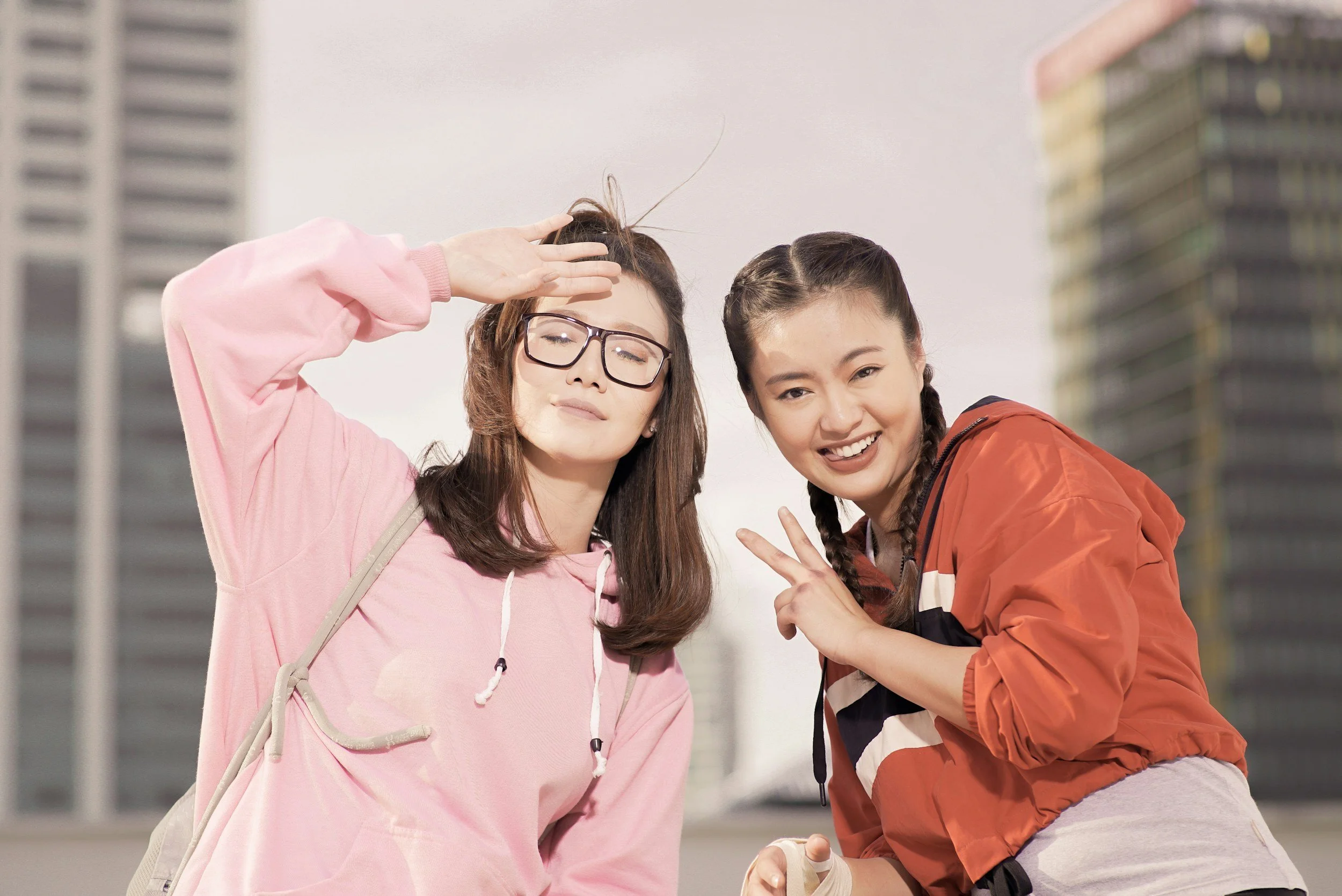 Two young women smiling and posing for a photo outdoors in an urban area with tall buildings in the background. One woman is wearing glasses and a pink hoodie, while the other is in a red jacket and making a peace sign.