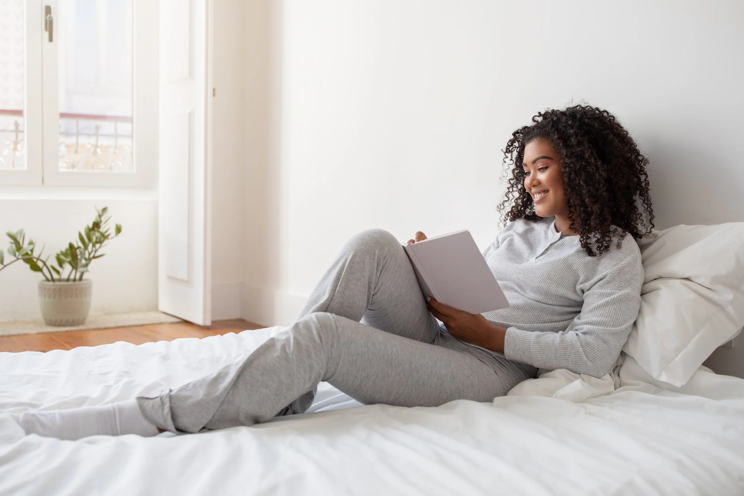 Young woman with curly hair smiling and reading a book while lying on a bed in a bright room.