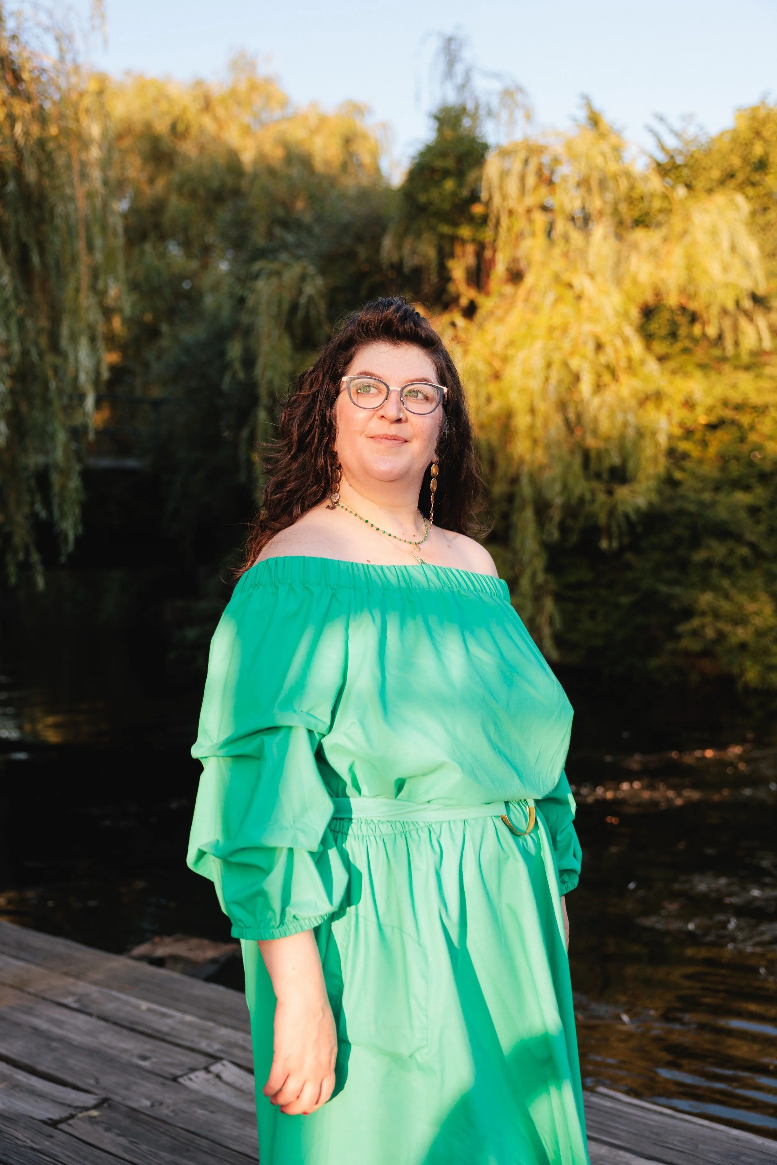 A woman with brown curly hair, glasses, and earrings, standing on a wooden dock near a river with trees and autumn foliage in the background, wearing a bright green off-the-shoulder dress.