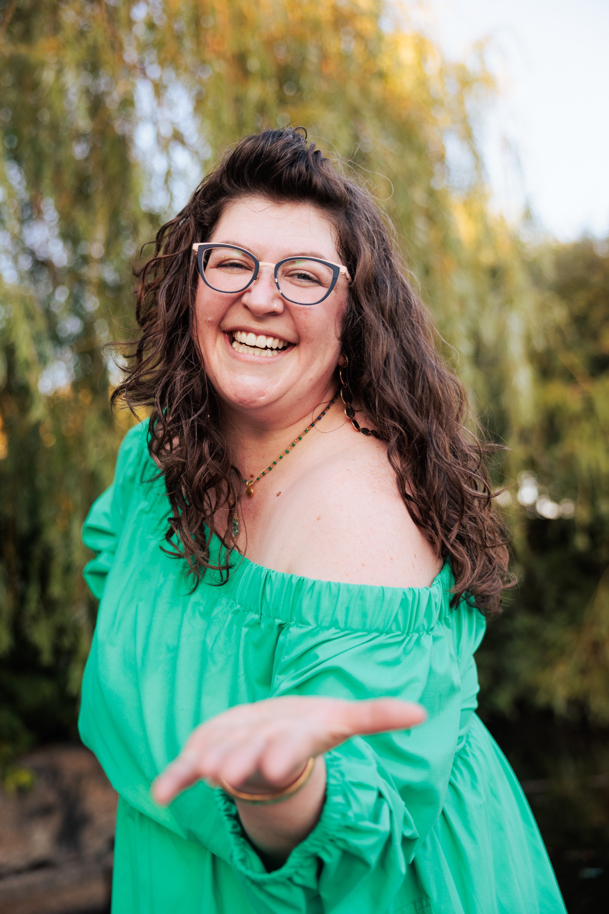 A woman with curly brown hair, glasses, and a nose ring, smiling and looking at the camera outdoors during fall, wearing a bright green off-shoulder dress and jewelry, holding out a hand invitingly, with trees and yellow leaves in the background.