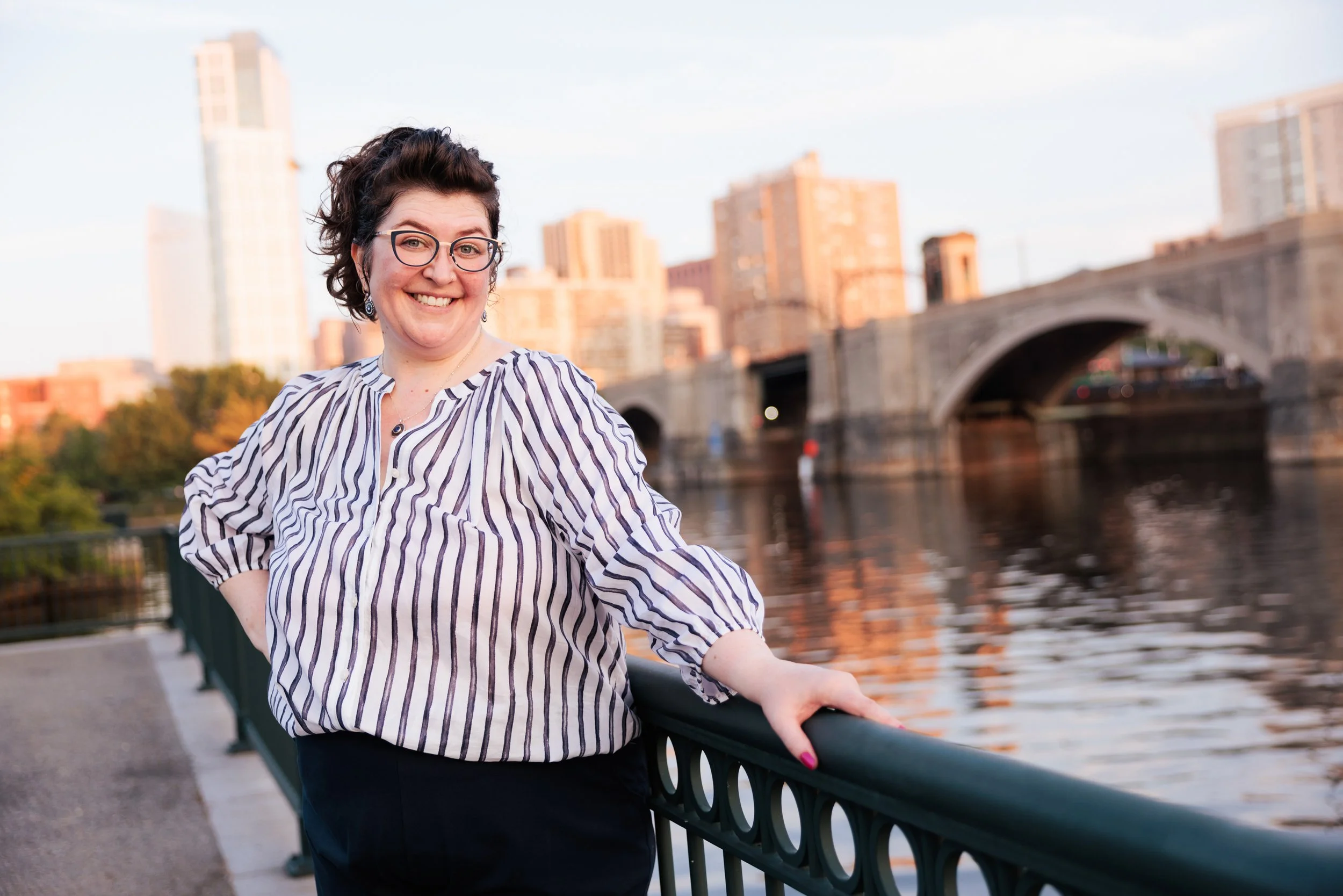 A woman with glasses and curly dark hair smiling, standing by a river with a cityscape and bridge in the background during sunset.