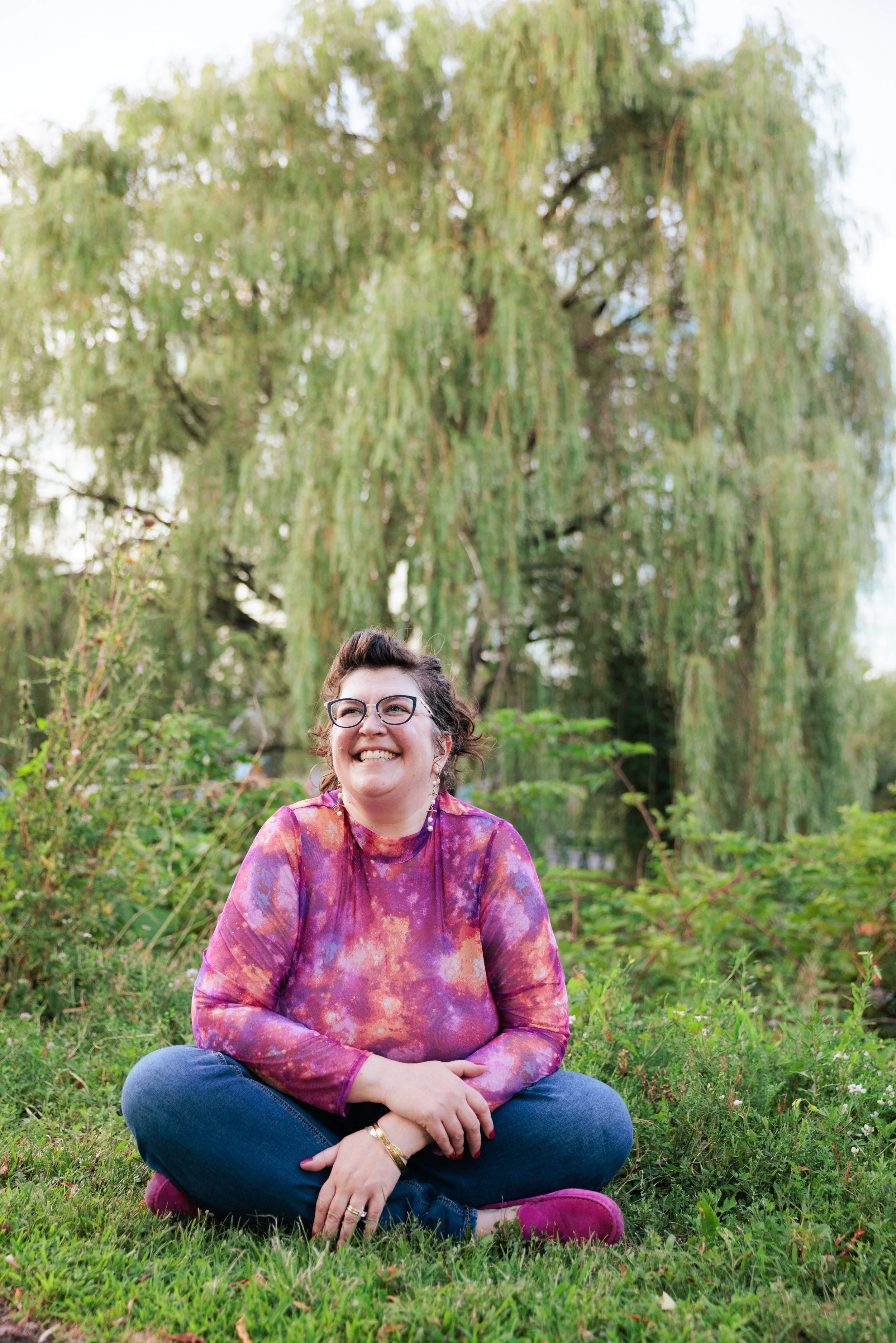 A woman sitting cross-legged on grass, smiling, with large trees in the background.