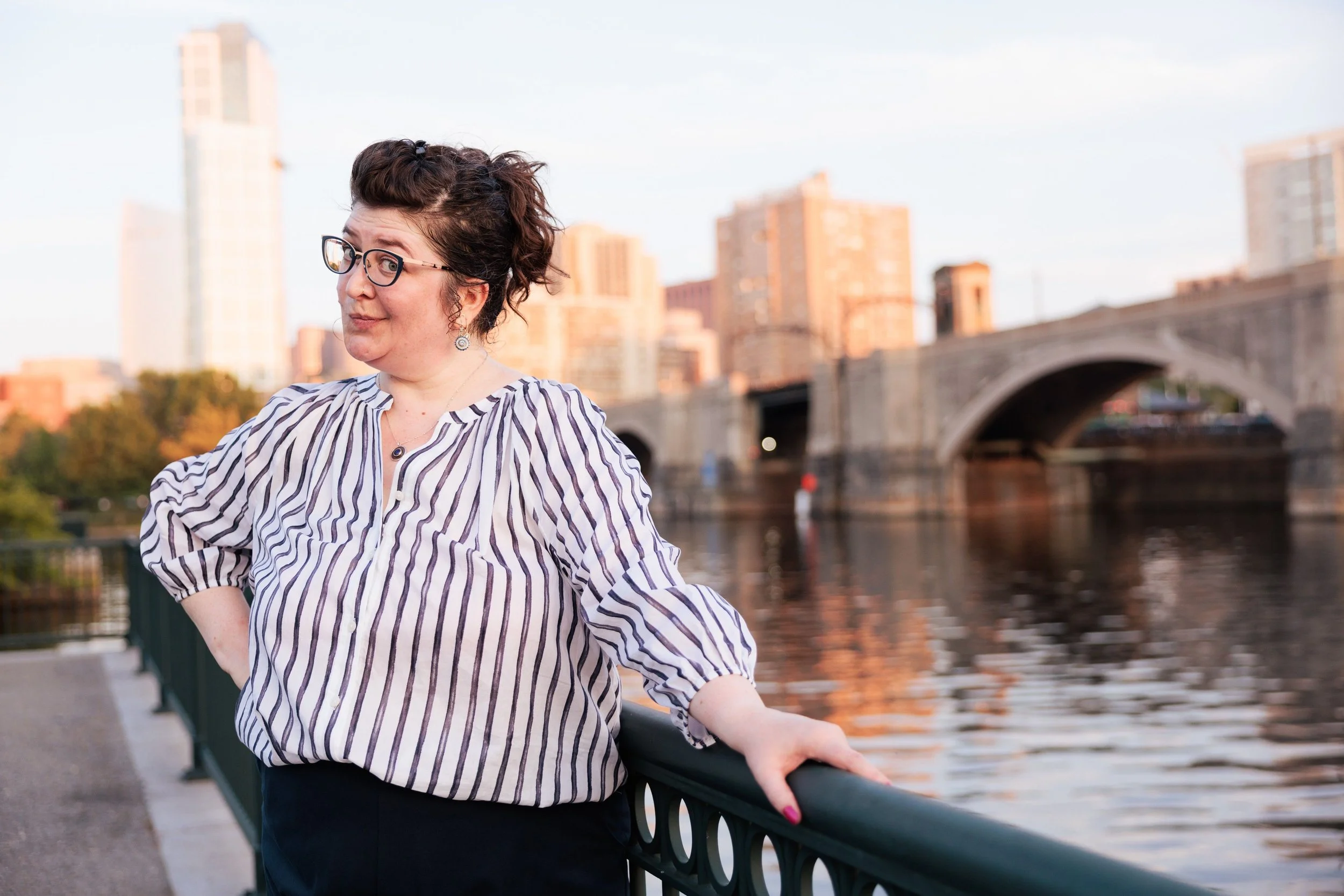 A woman with glasses, dark curly hair, and wearing a striped blouse stands by a river with a city skyline and bridge in the background during sunset.