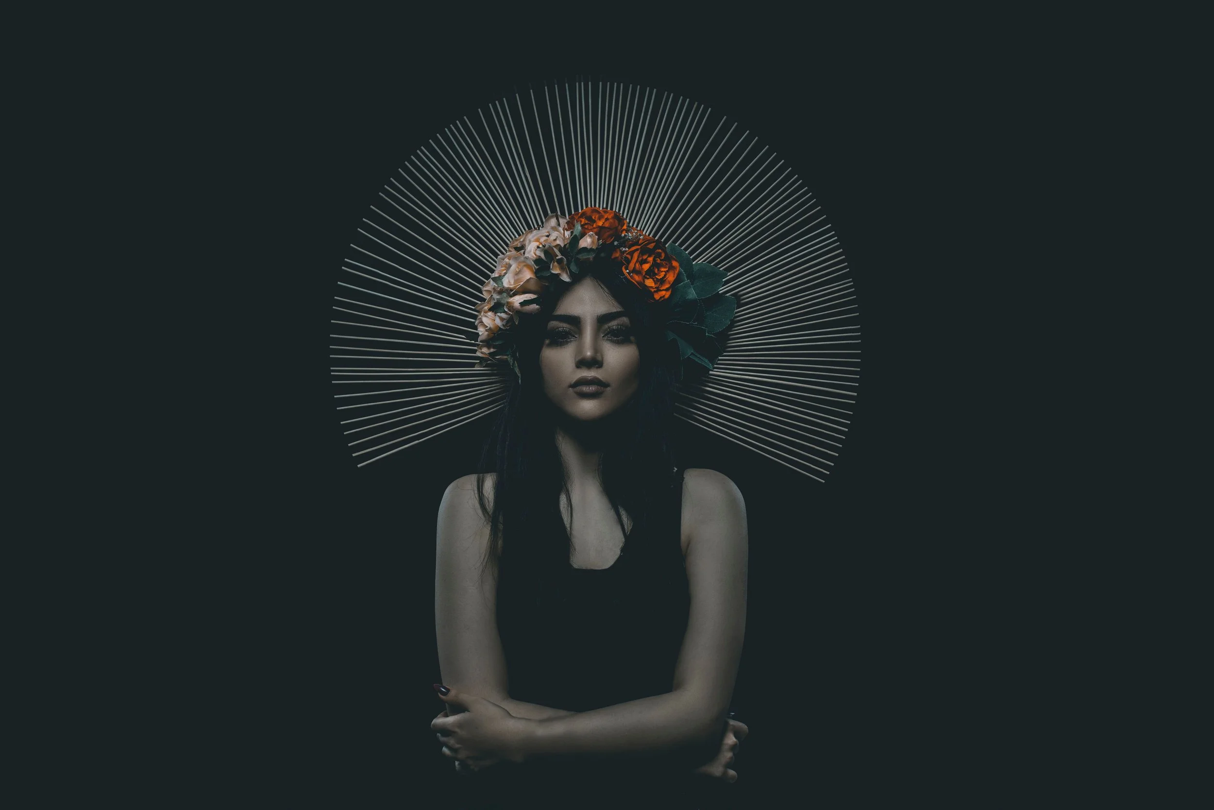 A woman with dark hair and makeup, wearing a floral headpiece, stands against a dark background with radiating lines behind her.