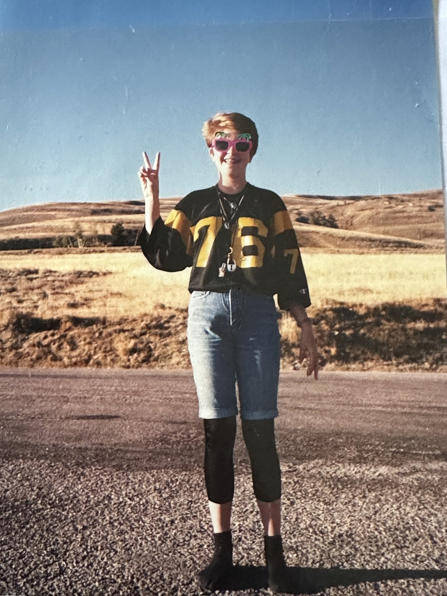 Jackie standing on the side of a road on a sunny day in spring giving a peace sign, a big smile, with sunglasses and an outfit only an 8th grader could love: leggings under jorts and a jr. high jersey.