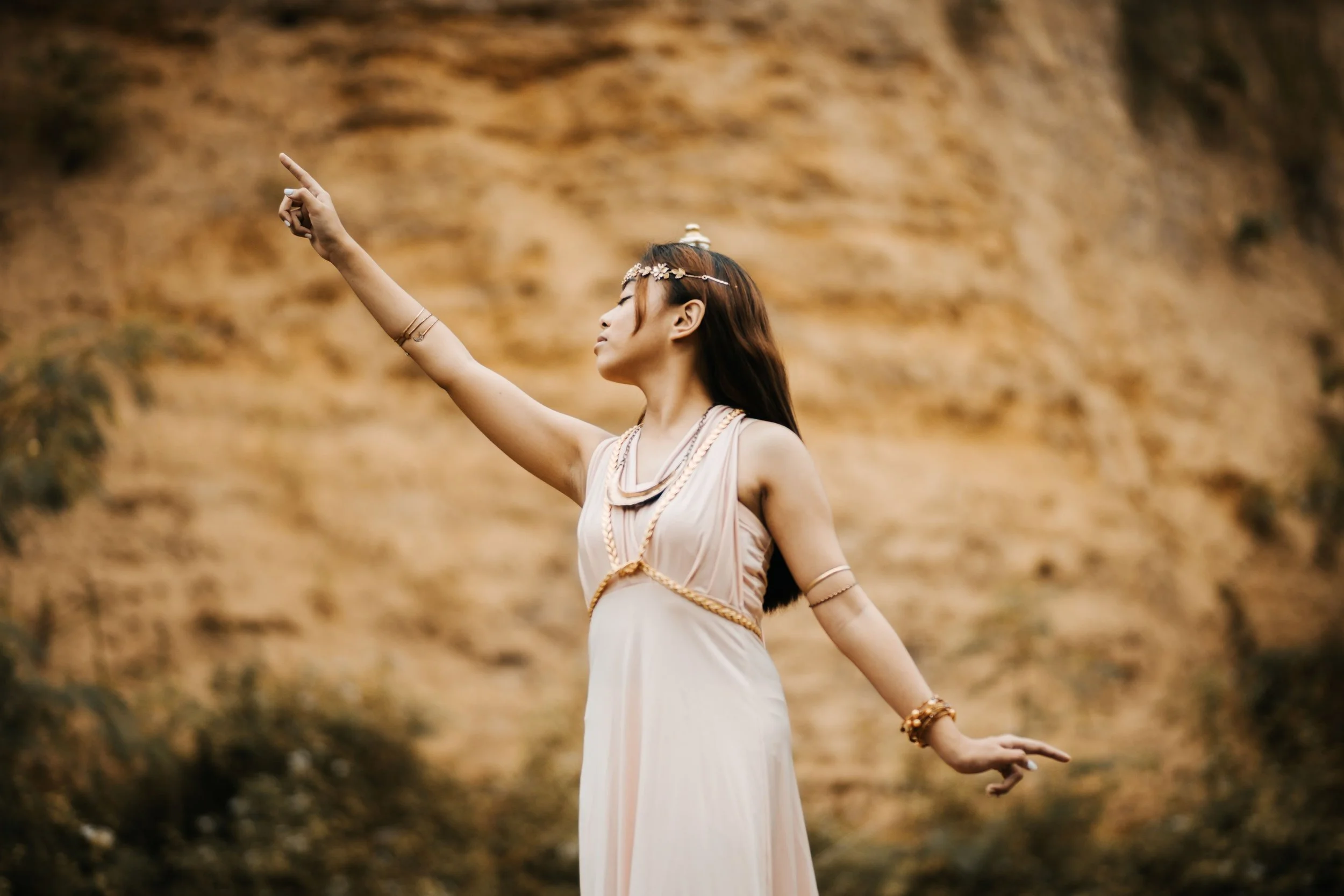A woman dressed in a flowing light-colored dress with accessories, wearing a floral headpiece, standing outdoors on a hillside with a blurred background of earthy rocks, with one arm extended and finger pointing upward.