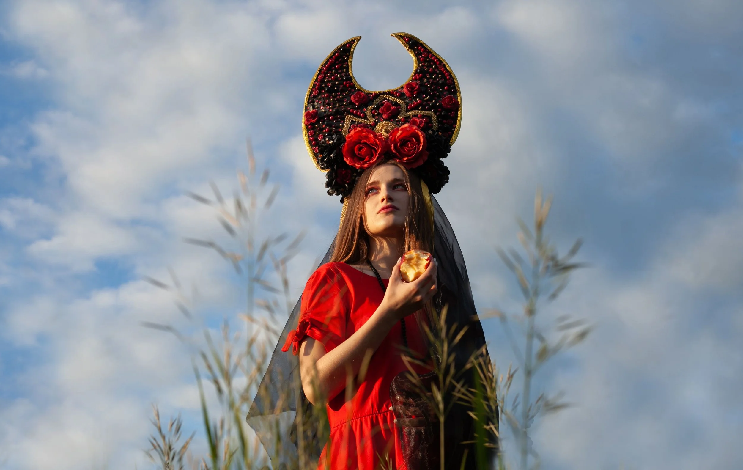 Young woman in a red dress and a large ornate headpiece holding a piece of bread, standing outdoors amid tall grass with a partly cloudy sky in the background.