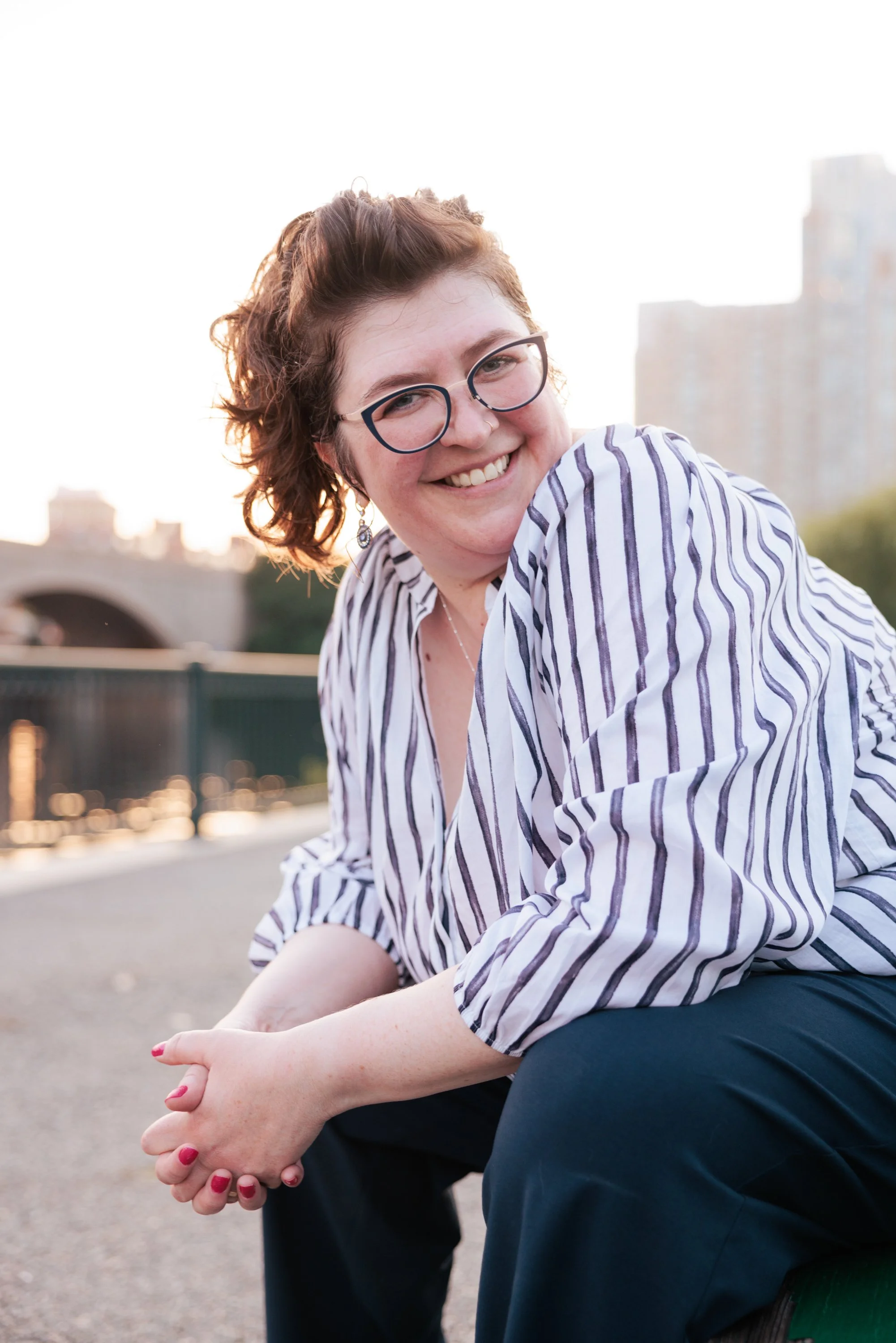 A woman with short curly hair, glasses, and earrings, smiling outdoors during sunset, wearing a white and navy striped shirt and navy pants, sitting near a bridge with city buildings in the background.