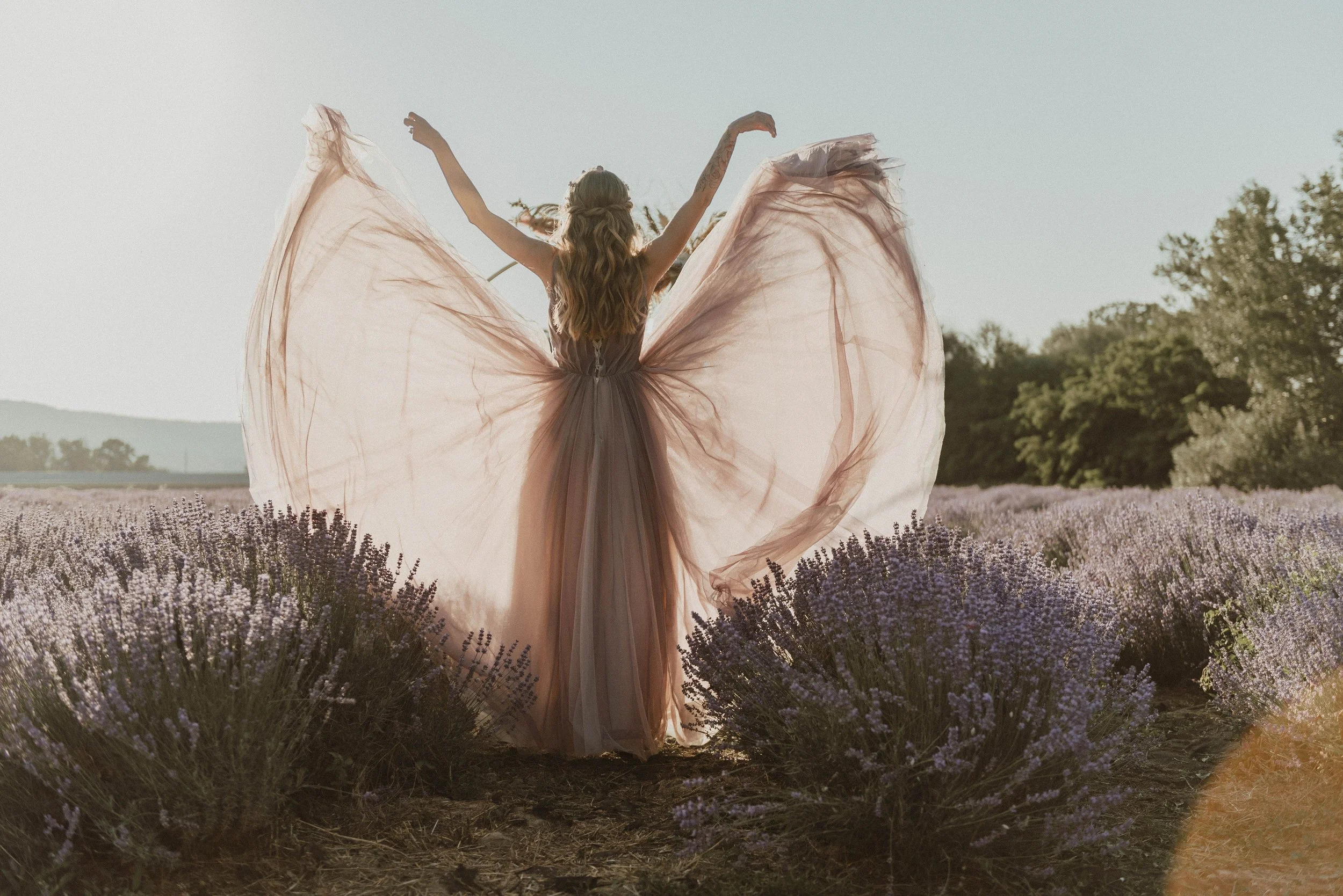 A woman wearing a flowing pink dress stands in a lavender field with her arms raised, enjoying the outdoors during daylight.