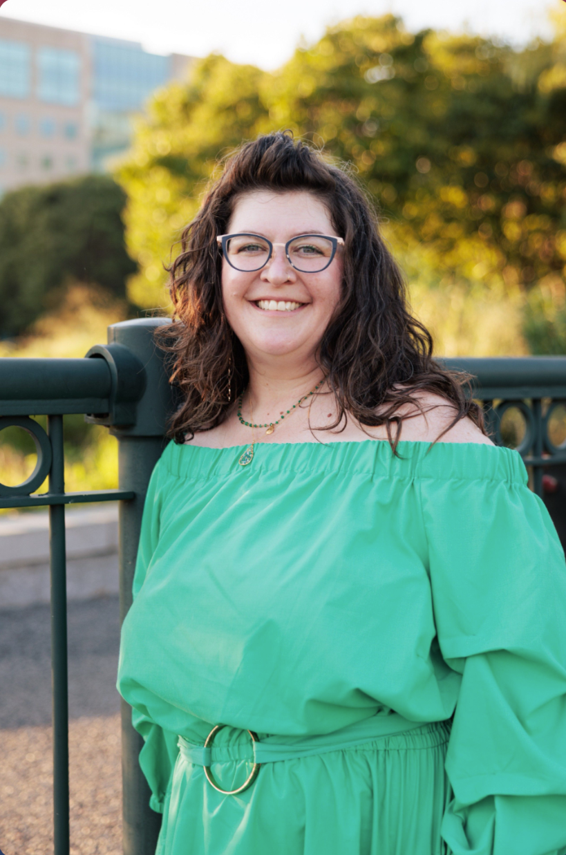 A woman with curly dark hair, glasses, and a bright smile, wearing an off-the-shoulder green dress, standing outdoors near a green railing with trees and a cityscape in the background during late afternoon or early evening.