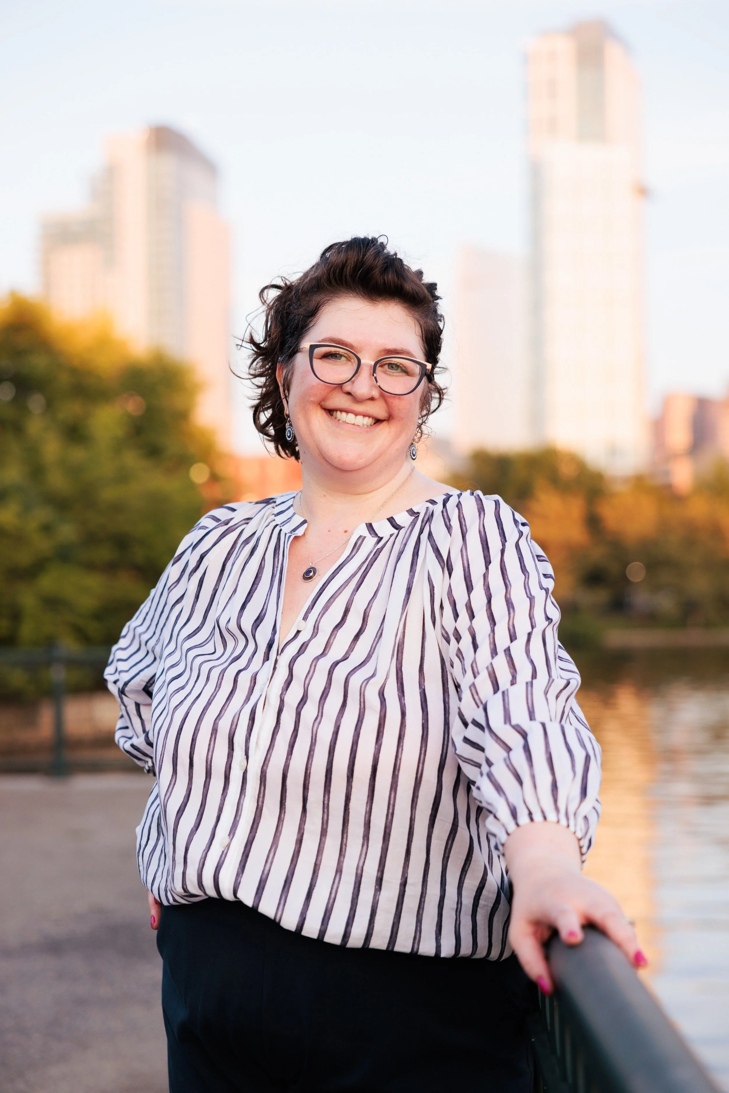 Jackie smiling and looking at the camera in a white and navy striped shirt and navy pants with an arm resting on a handrail on the river in a park with a city scape in the background.