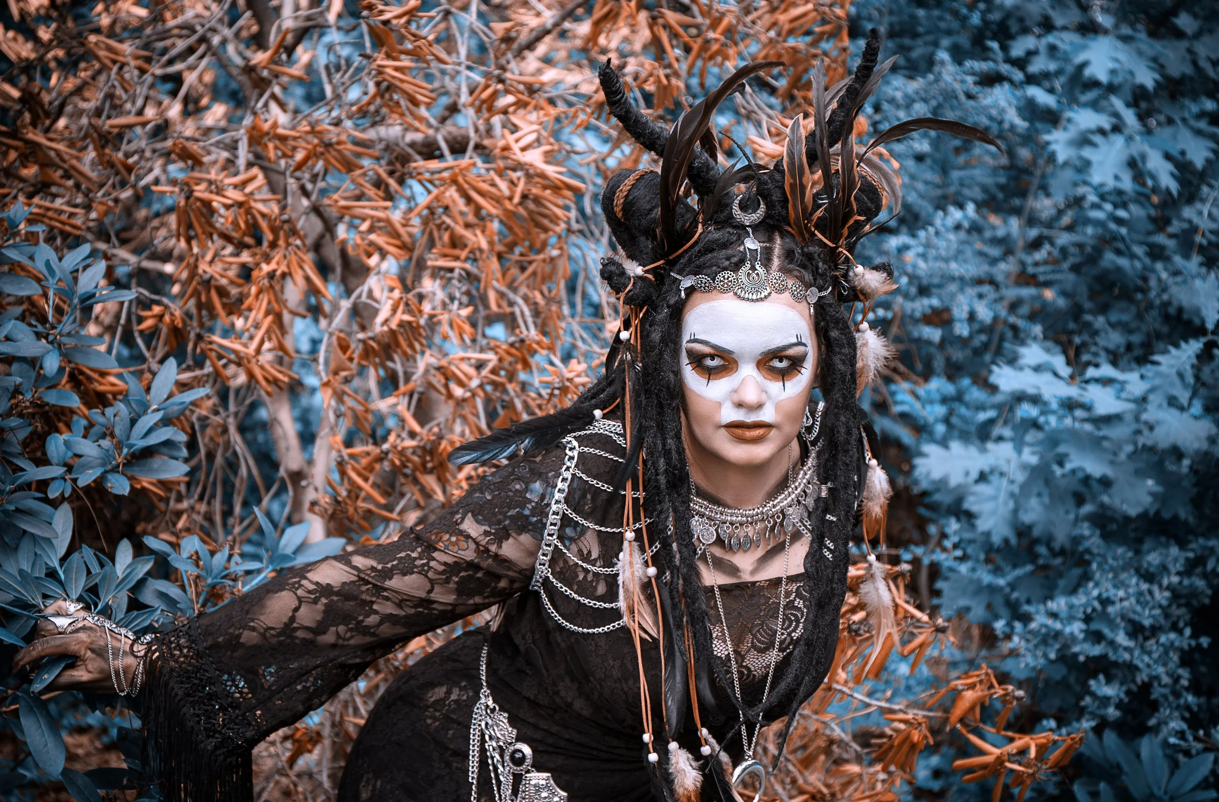 A woman dressed in a costume with a tribal or fantasy theme, wearing face paint resembling a skull, a headdress with feathers, jewelry, and dark clothing, standing outdoors among orange and blue-tinted foliage.