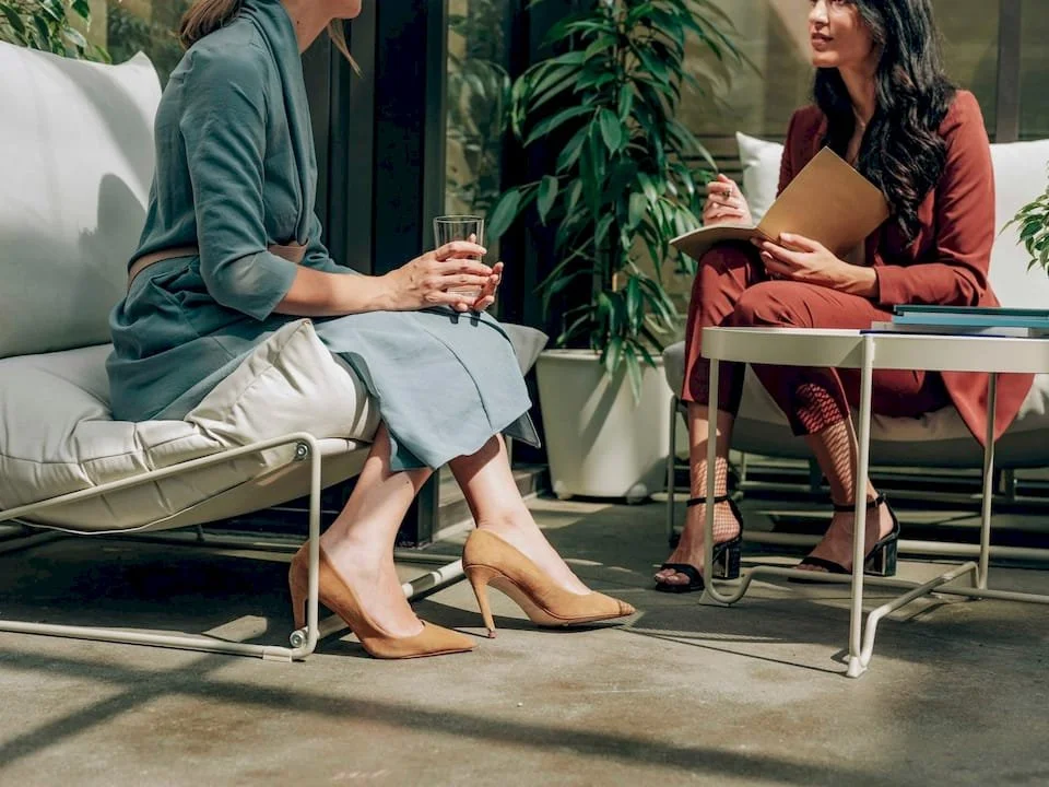 Two women speaking in formal business attire, outside at a therapy session. One is wearing heels and facing each other.