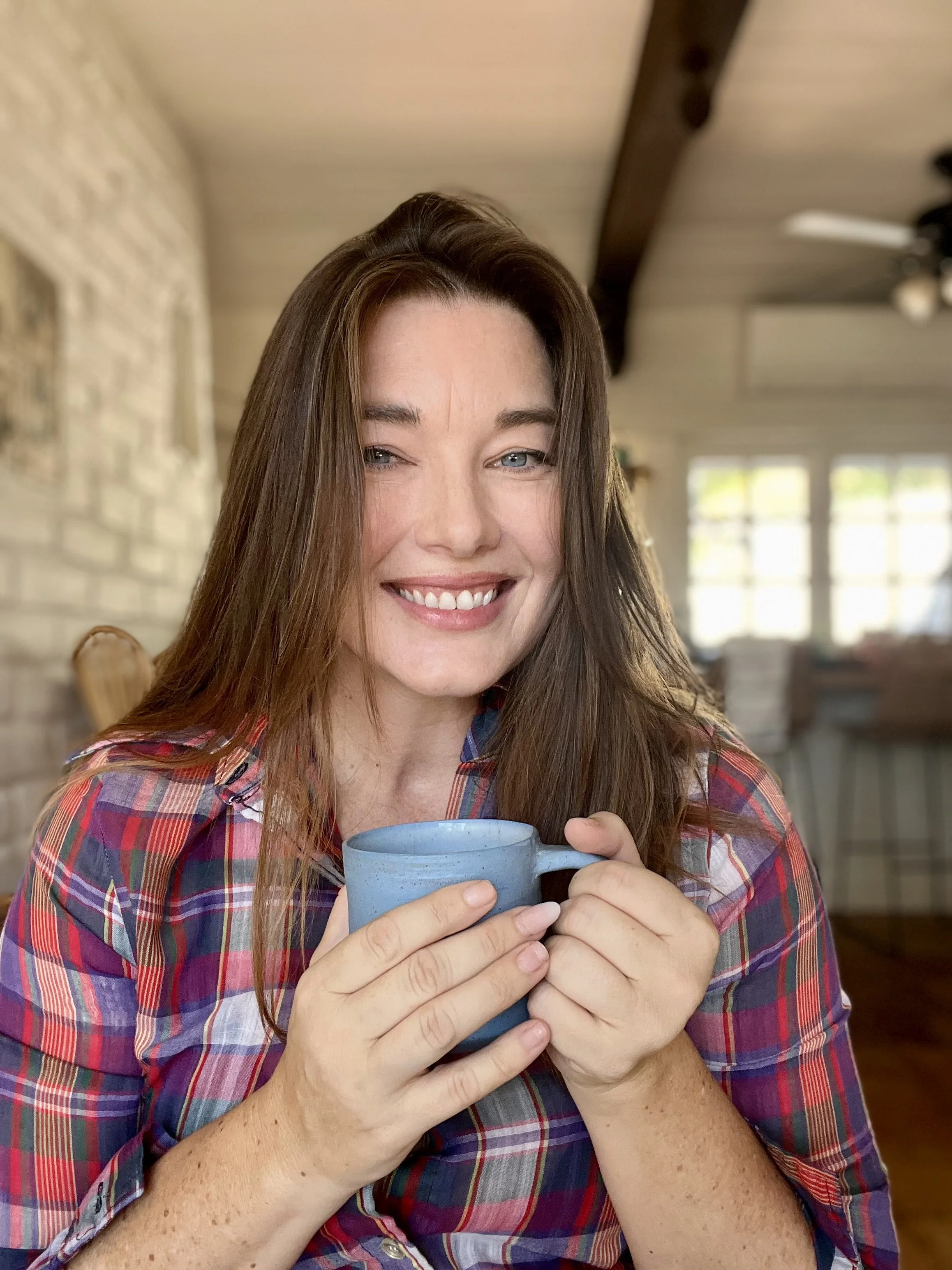 A woman with long brown hair smiling and holding a blue coffee mug in a cozy, well-lit room with large windows, beige walls, and wooden ceiling beams.