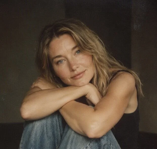 A woman with wavy blonde hair resting her head on her crossed arms, smiling gently at the camera.