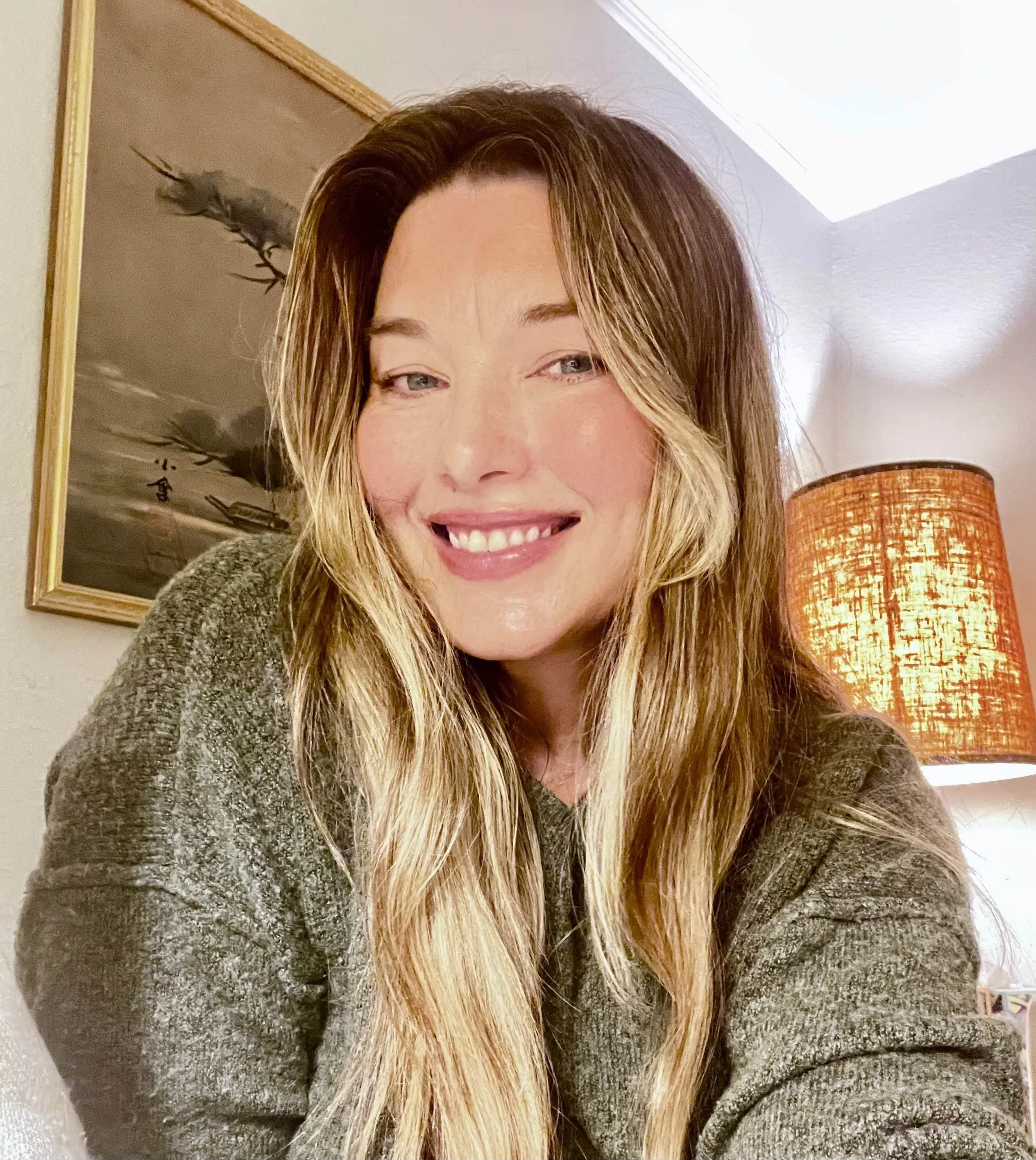 Kelli Redfield, LMFT with long blonde hair and light skin smiling in a cozy room. Behind her, there is a framed artwork of flying birds and a table lamp with a textured orange lampshade. The ceiling is white with a part of a skylight visible.