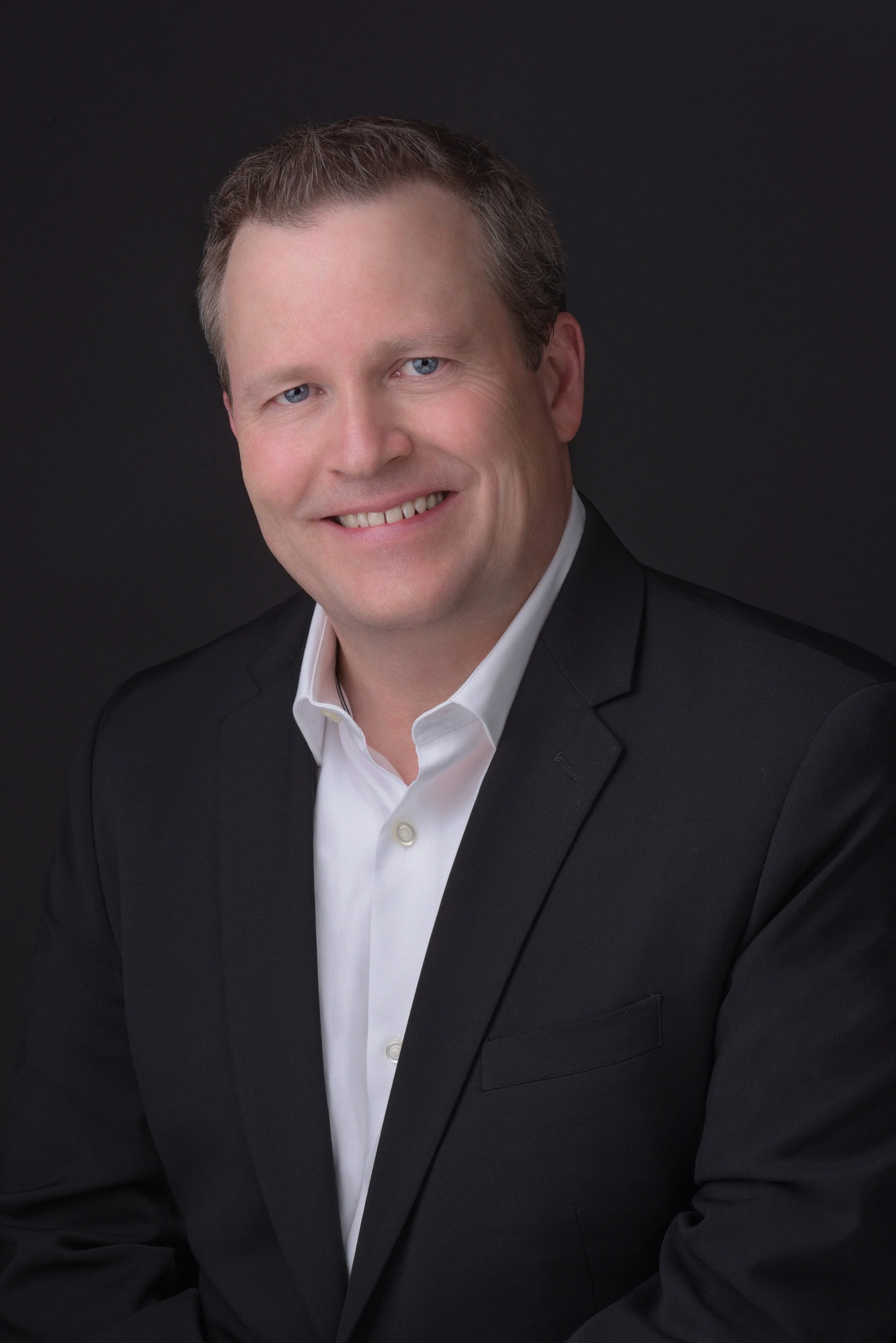 Professional headshot of a smiling man with light brown hair, wearing a black suit and white shirt, against a dark background.