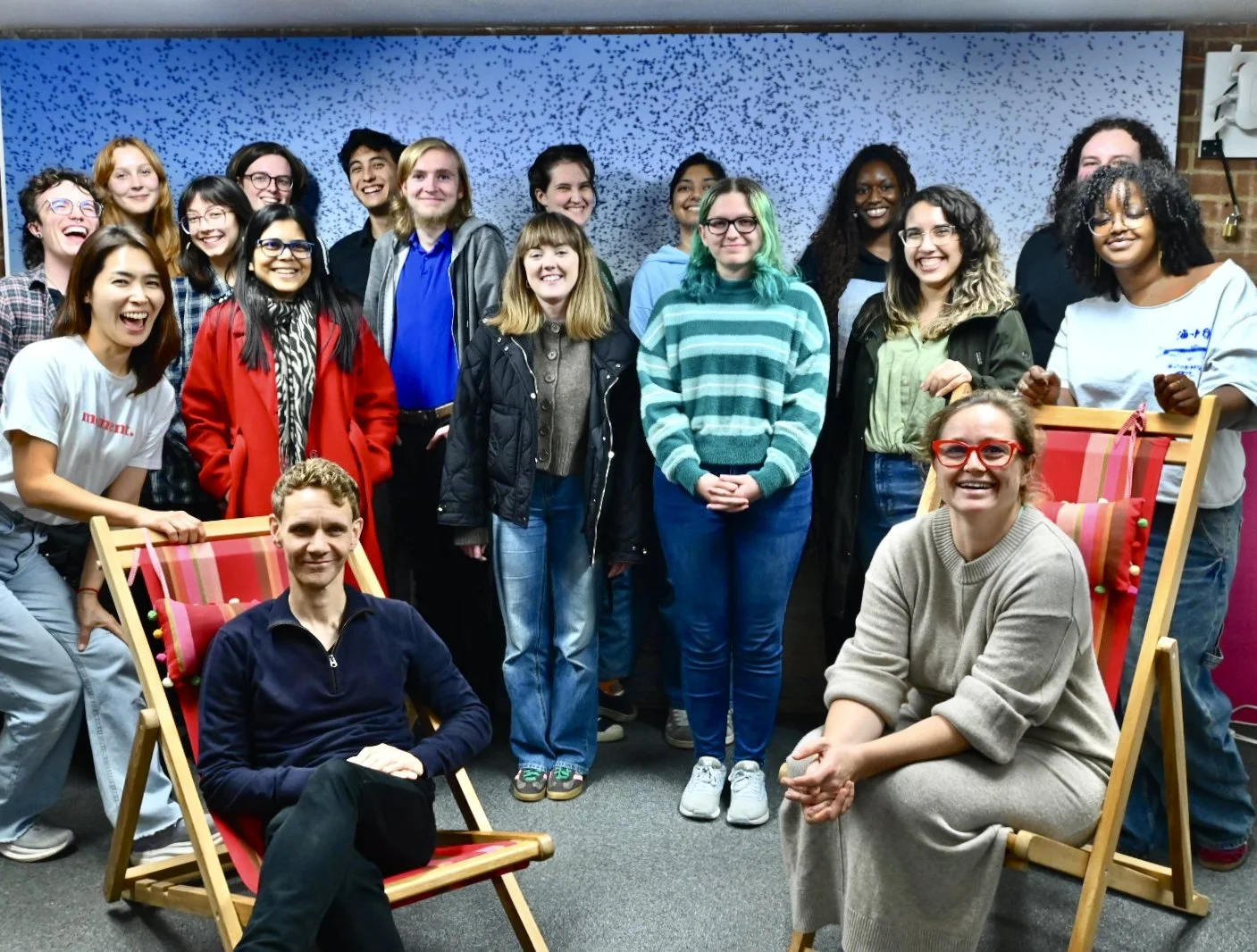 A diverse group of people, including men and women, are standing and sitting in an indoor setting, smiling and posing for a group photo. The background features a blue and black speckled wall and a brick wall on the right.