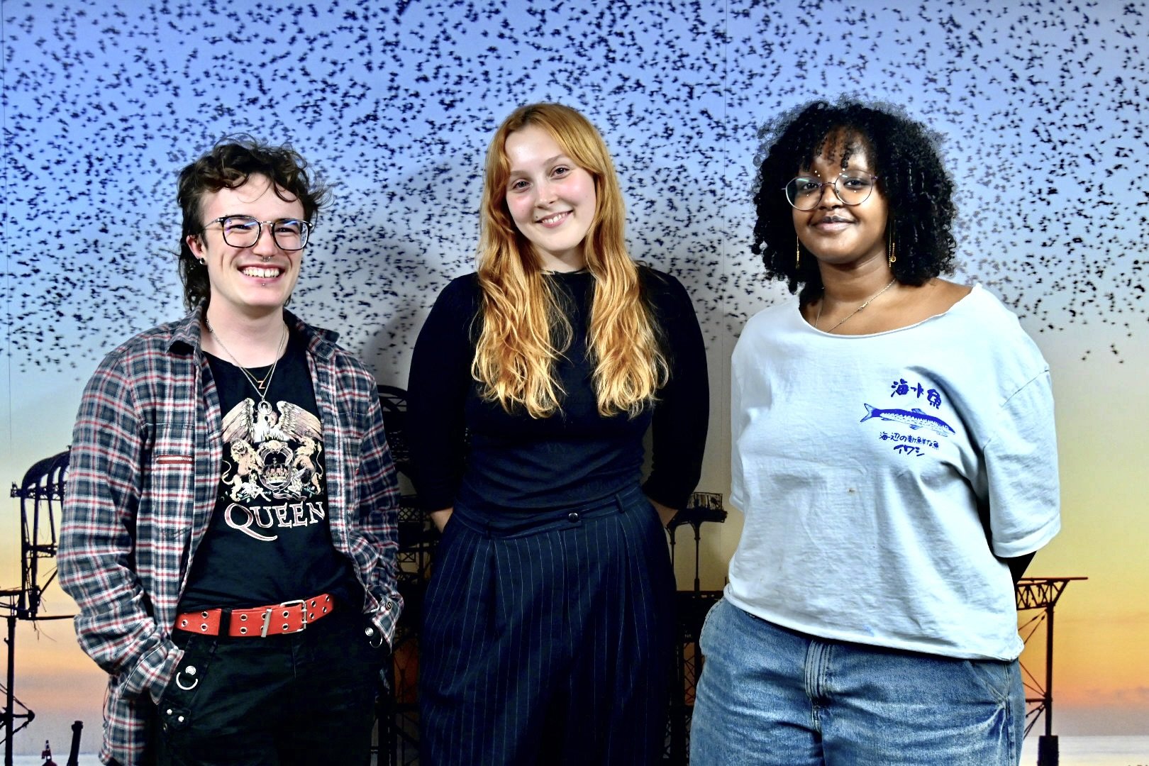 Three women standing outdoors with a backdrop of birds flying in the sky at sunset, forming a silhouette of structures.