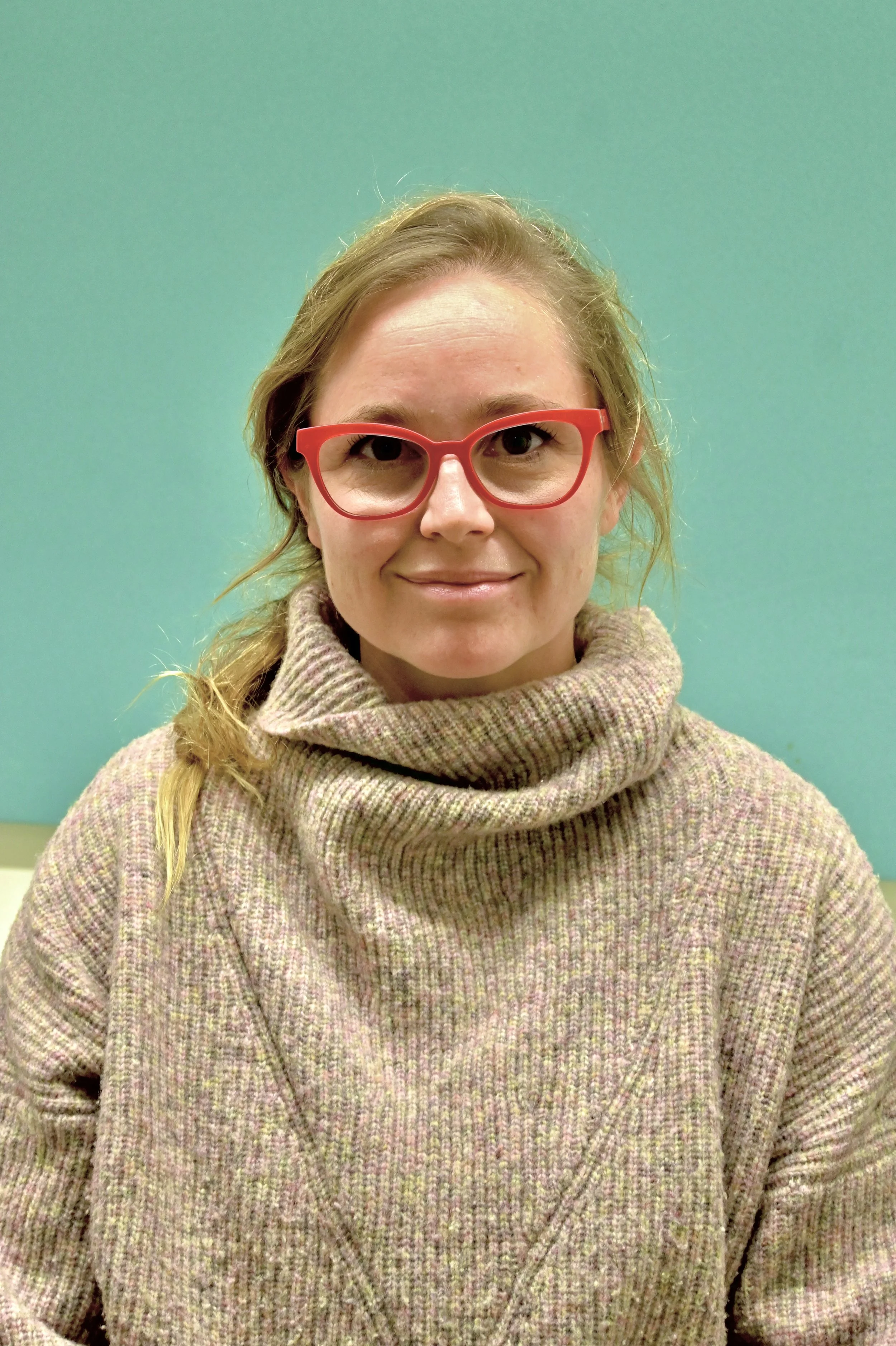 A woman with blonde hair, wearing red glasses and a turtleneck sweater, sitting in front of a teal wall.