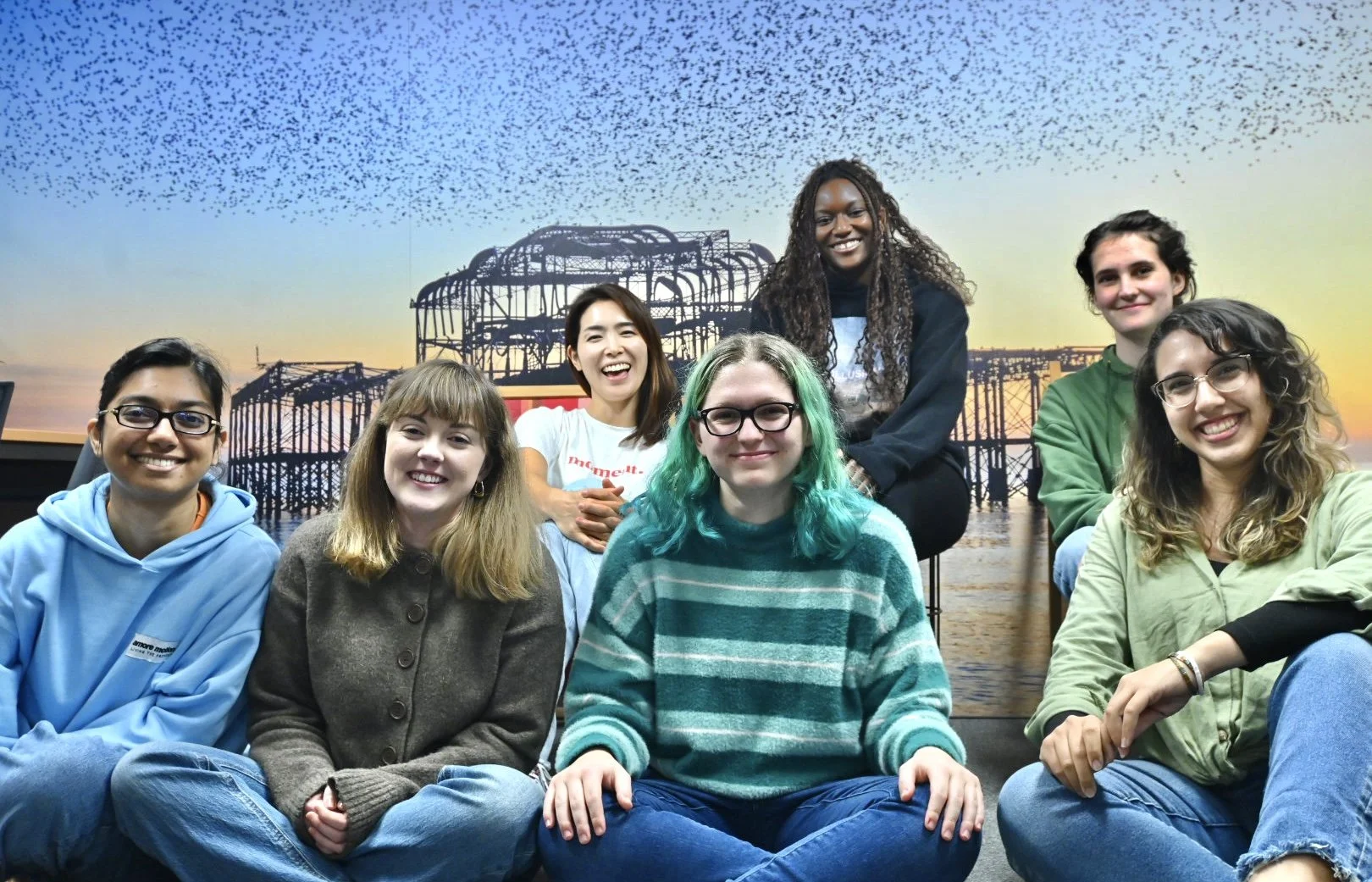 Group of eight diverse young women smiling and posing for a photo indoors with a scenic sunset and architectural background mural.