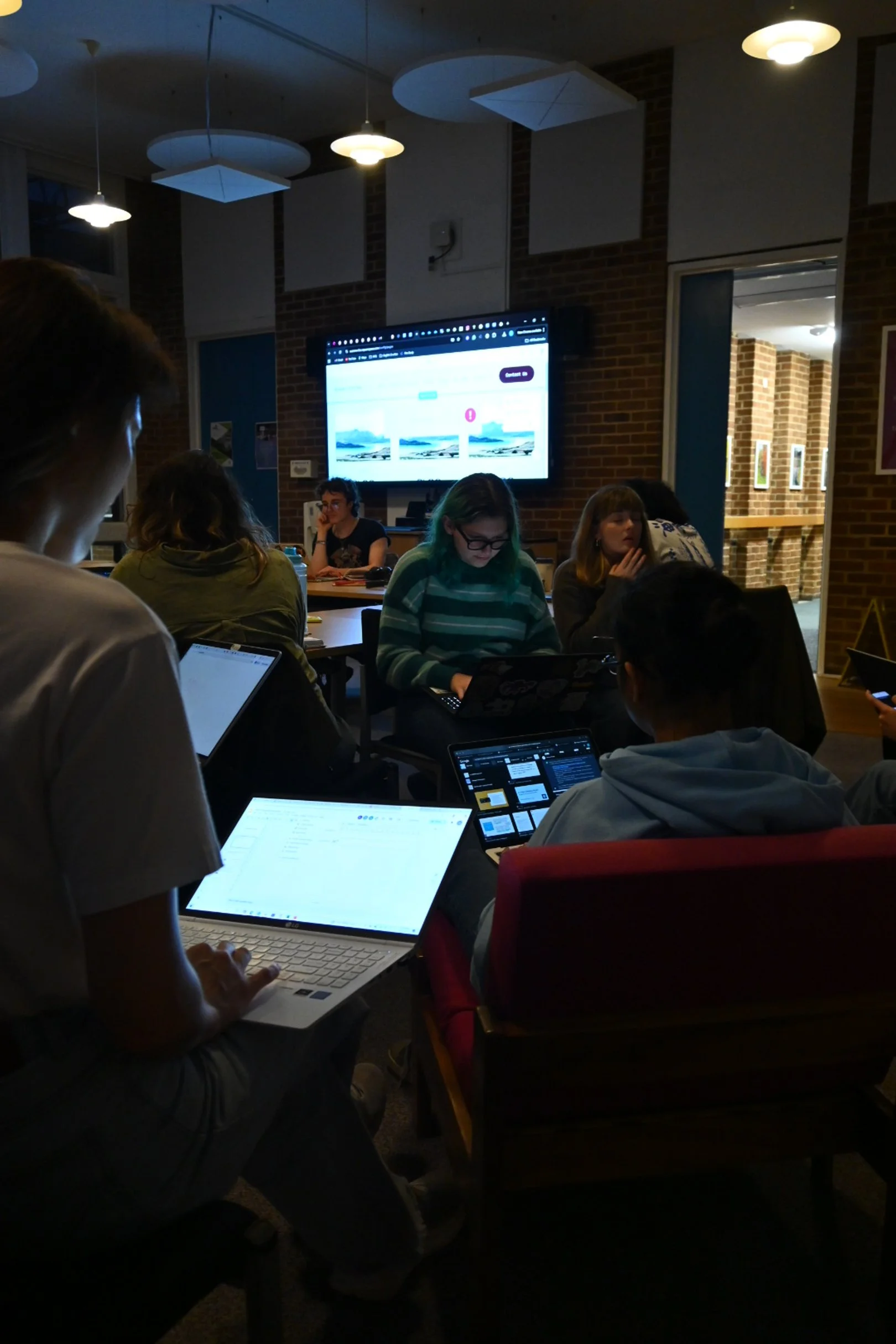 People seated around a table in a dimly lit room, working on laptops and tablets, with a large screen displaying a website in the background.