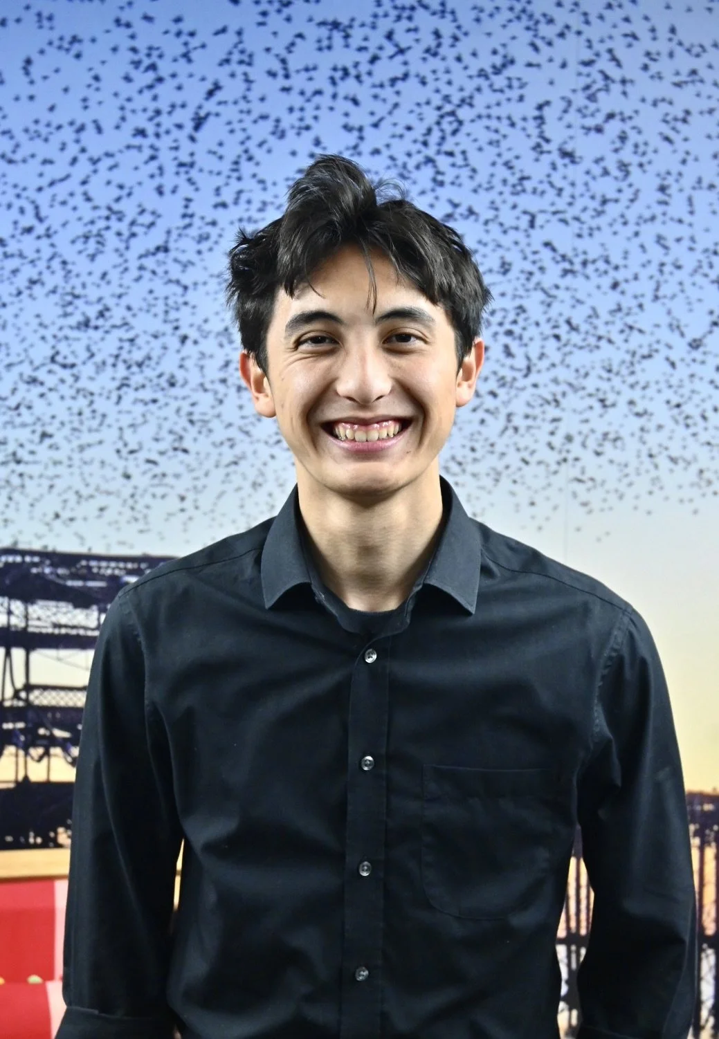 A young man with short dark hair, smiling, wearing a black button-up shirt, standing in front of a sunset sky with a large flock of birds flying and a bridge in the background.