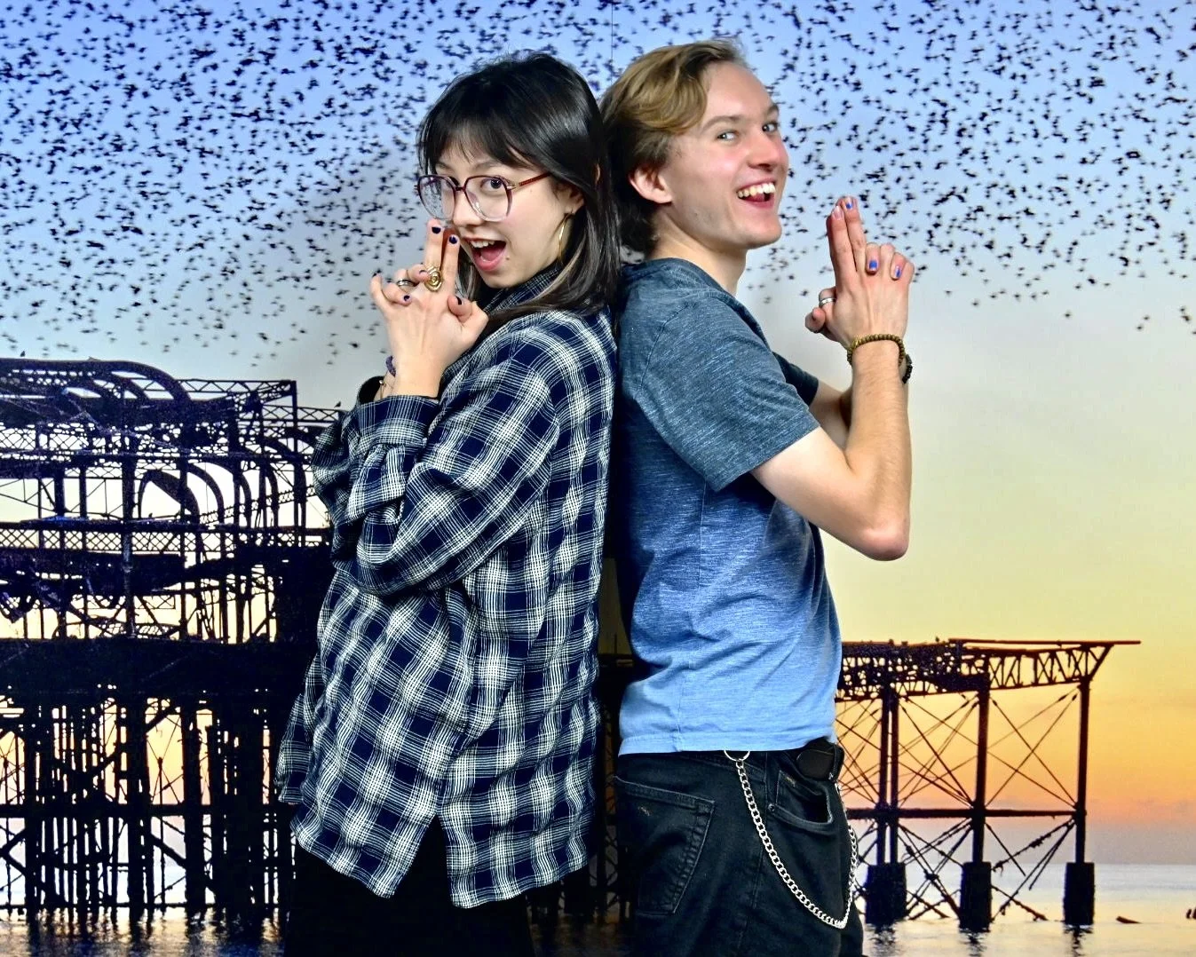 Two young adults standing back-to-back on a pier at sunset, smiling and making playful hand gestures, with a flock of birds flying overhead and an industrial pier in the background.