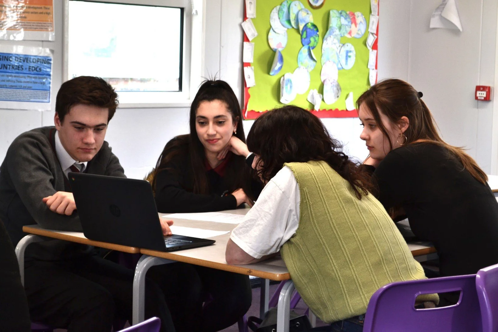 Four students sitting at a school table, looking at a laptop, engaging in a group activity or discussion.