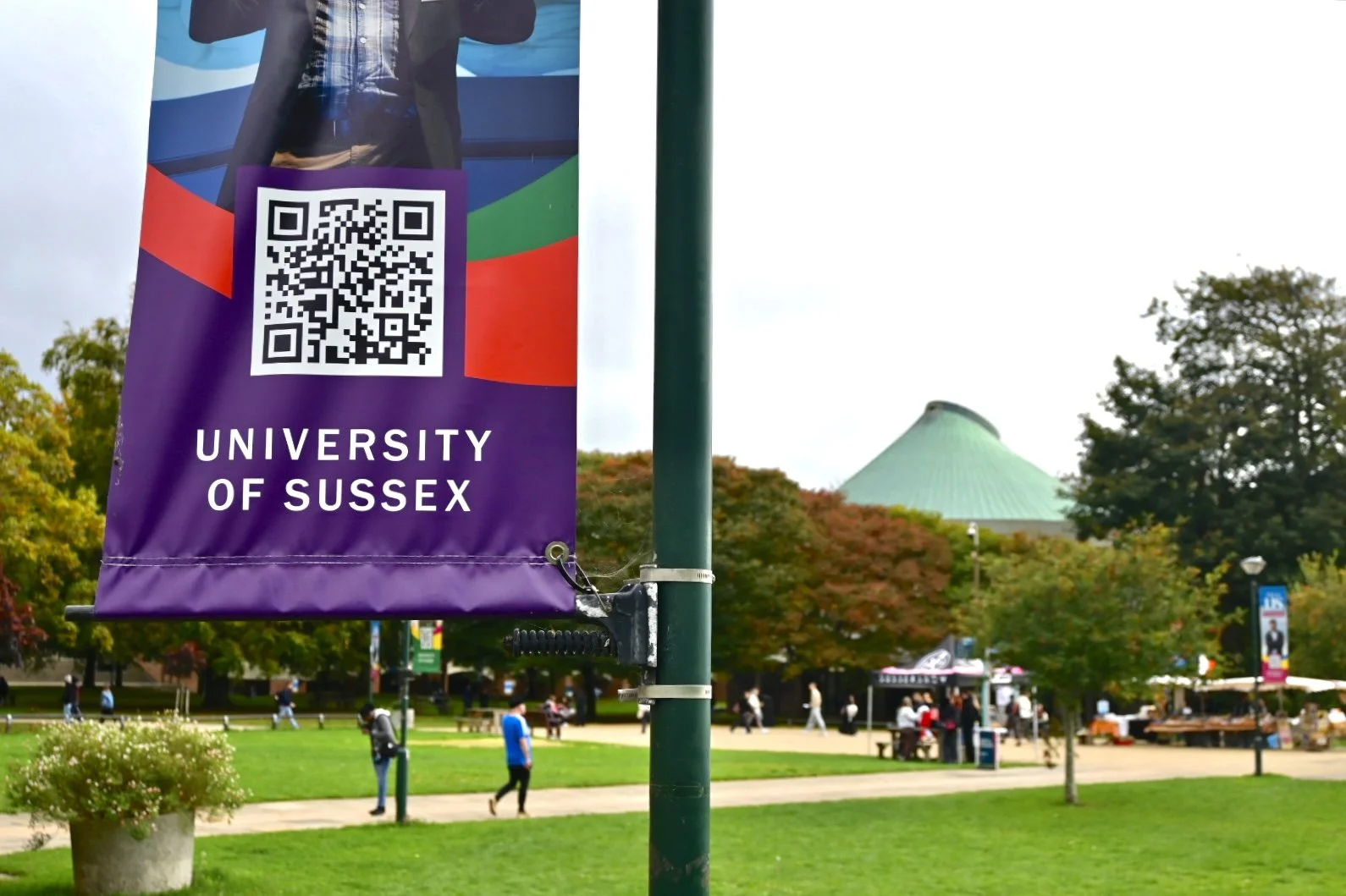 A university banner for the University of Sussex hanging on a lamppost, with a park scene in the background including trees, people walking, and a structure with a curved green roof.