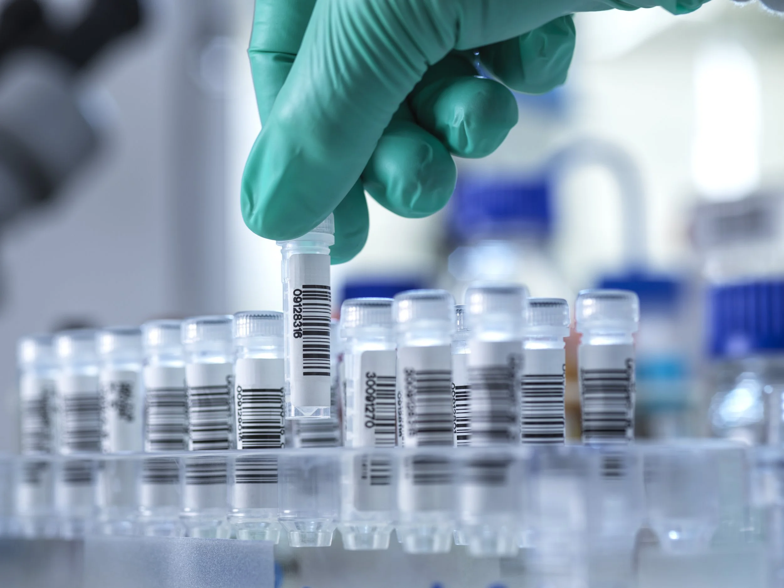 A gloved hand placing a vial with barcode labels into a rack of similar vials in a laboratory setting.