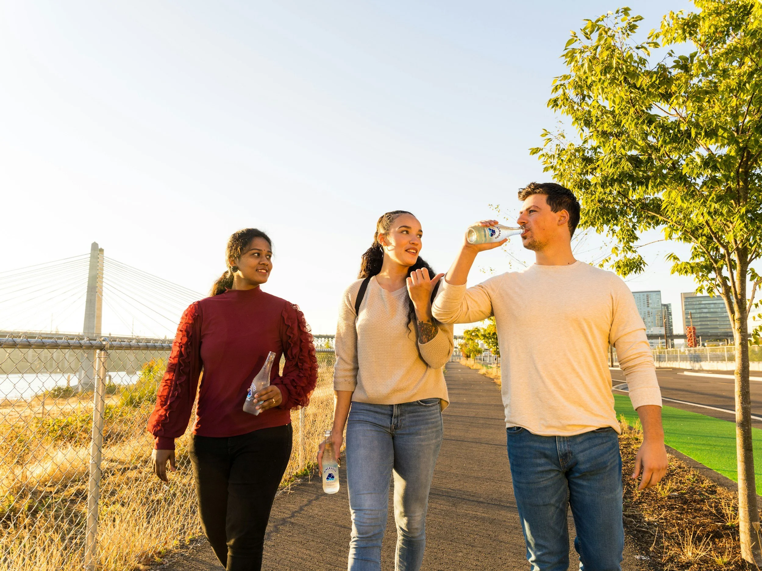 Three young adults walking outdoors near a fence, with a bridge and city skyline in the background. The person on the right is drinking from a glass bottle, while the others are chatting.
