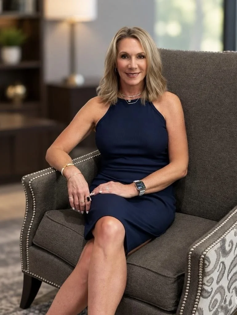 A woman with shoulder-length blonde hair wearing a navy blue sleeveless dress, sitting on a gray armchair with silver nailhead trim in a well-lit room with blurred background decor.
