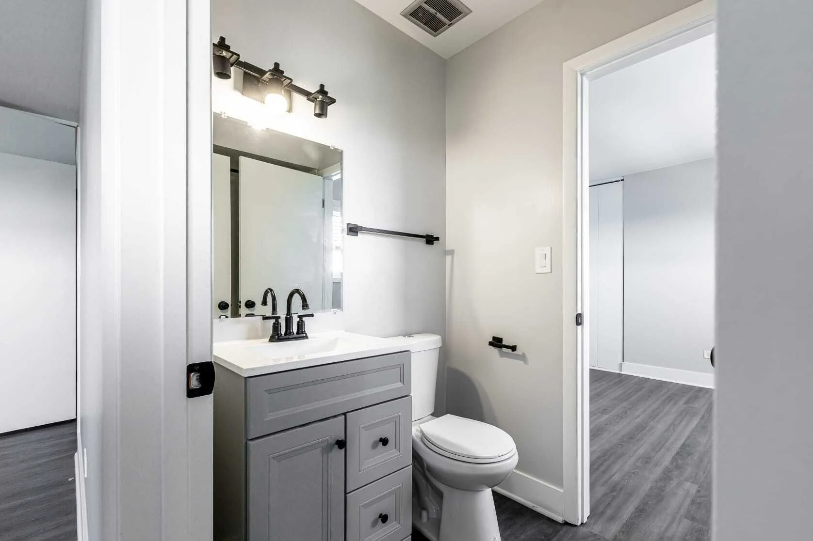 Bathroom with a sink vanity, mirror, toilet, and black fixtures, with a doorway leading to a room with grey walls and grey flooring.