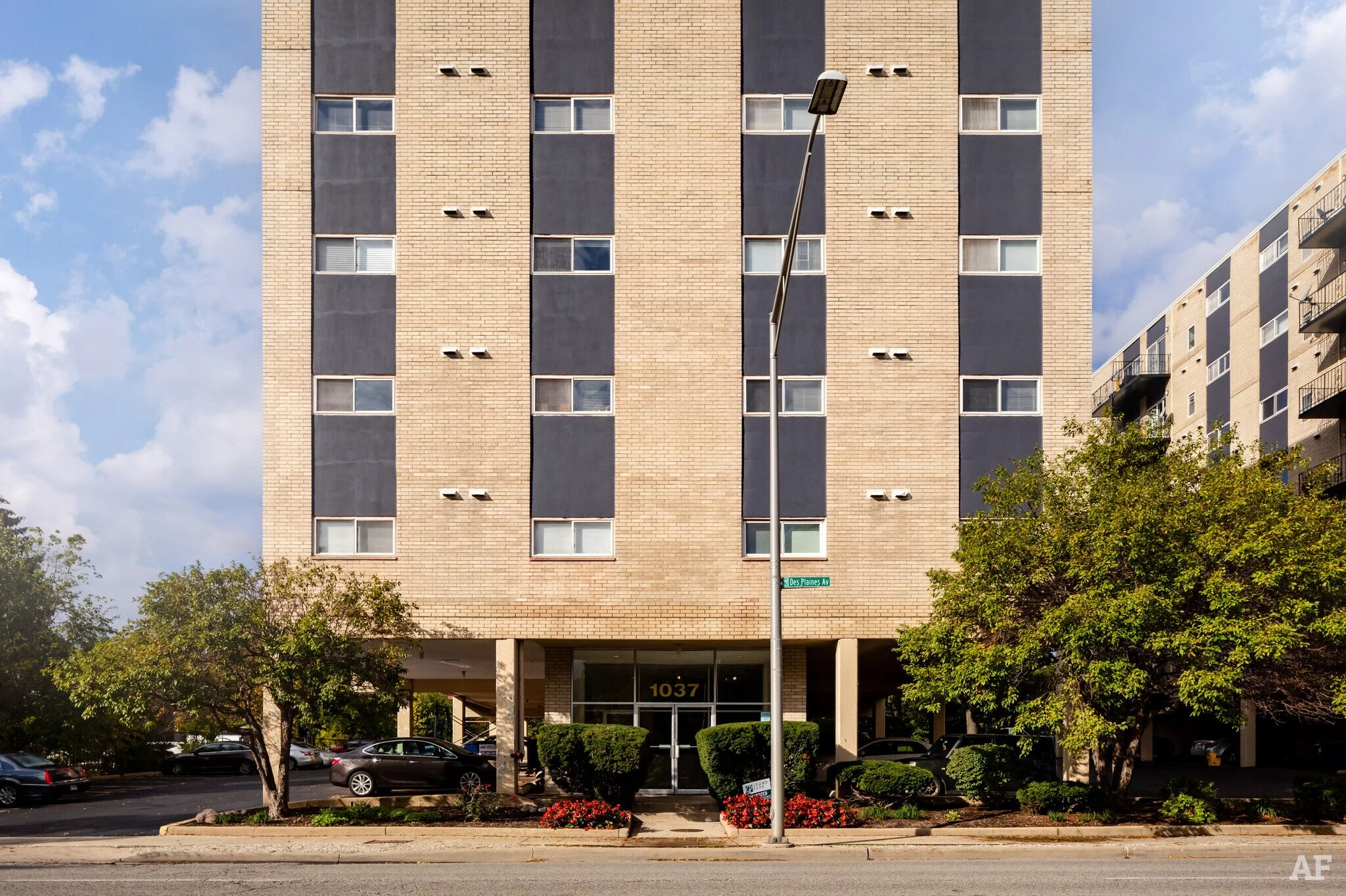 Front exterior of a multi-story residential apartment building with a brick facade, multiple windows, and a parking area underneath. Green trees and a streetlamp are in the foreground.