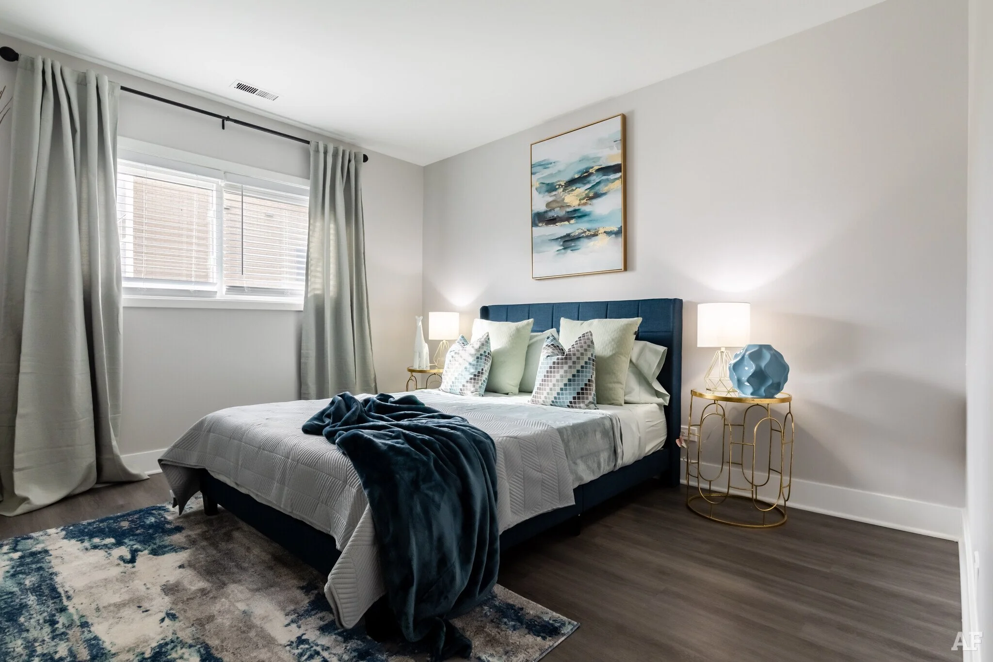 A modern bedroom with a bed, gray bedding, multiple pillows, two nightstands with lamps, a blue headboard, fresh curtains, a wall art, and a patterned rug.
