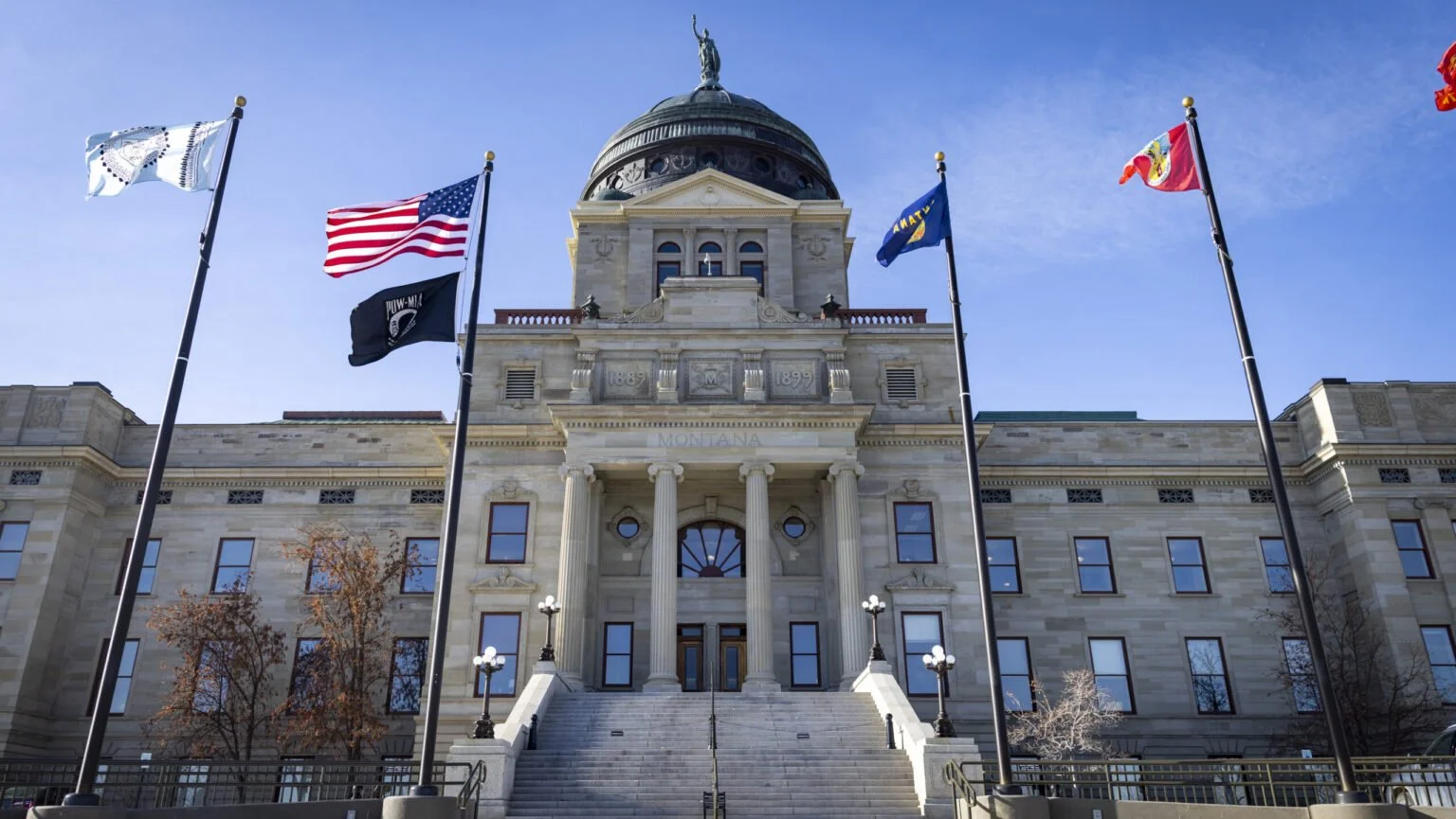 Historic government building with steps leading up to columns, surrounded by flagpoles with flags, under a blue sky.