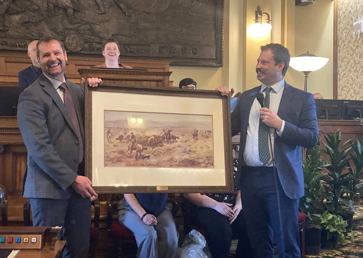 Two men in suits holding a framed picture of horses in a desert landscape, with one man speaking into a microphone as others look on, in a formal room with wood paneling and plants.