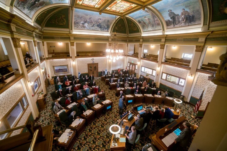 View of a legislative chamber with lawmakers seated at desks, large chandelier, ornate ceiling with painted murals, and American flag at the front.