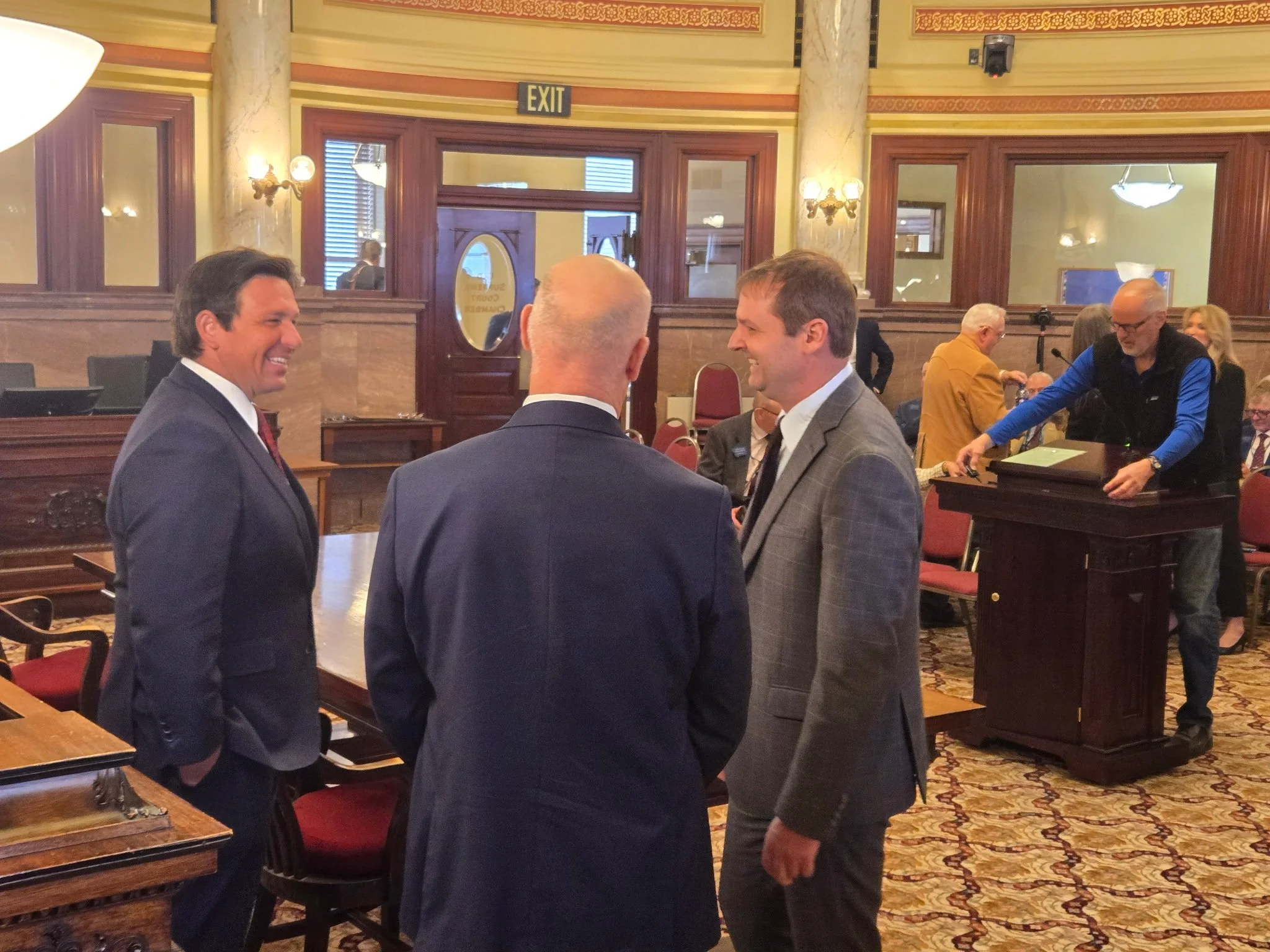 Three men in suits having a conversation in a wood-paneled room with ornate decor, red chairs, and a patterned carpet, while other people and a person at a podium are seen in the background.