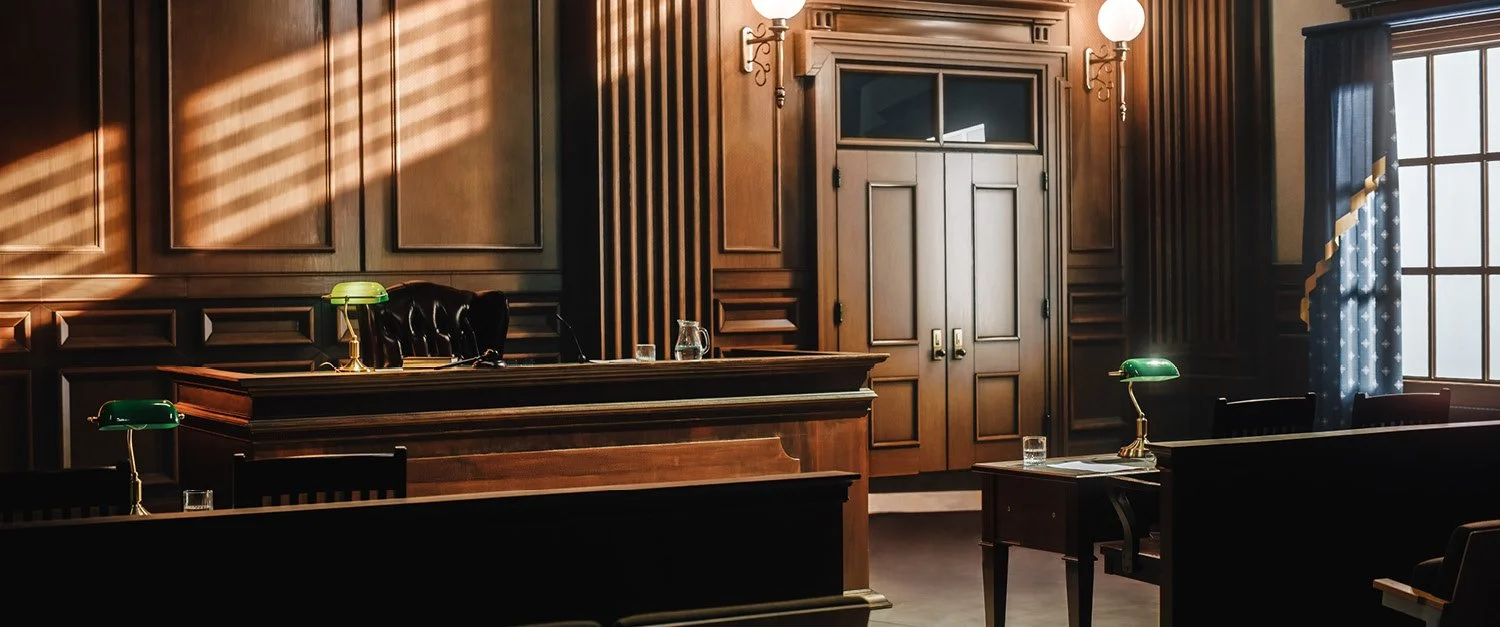 An empty courtroom with wooden paneling, green desk lamps, a judge's bench, and a large window with curtains.