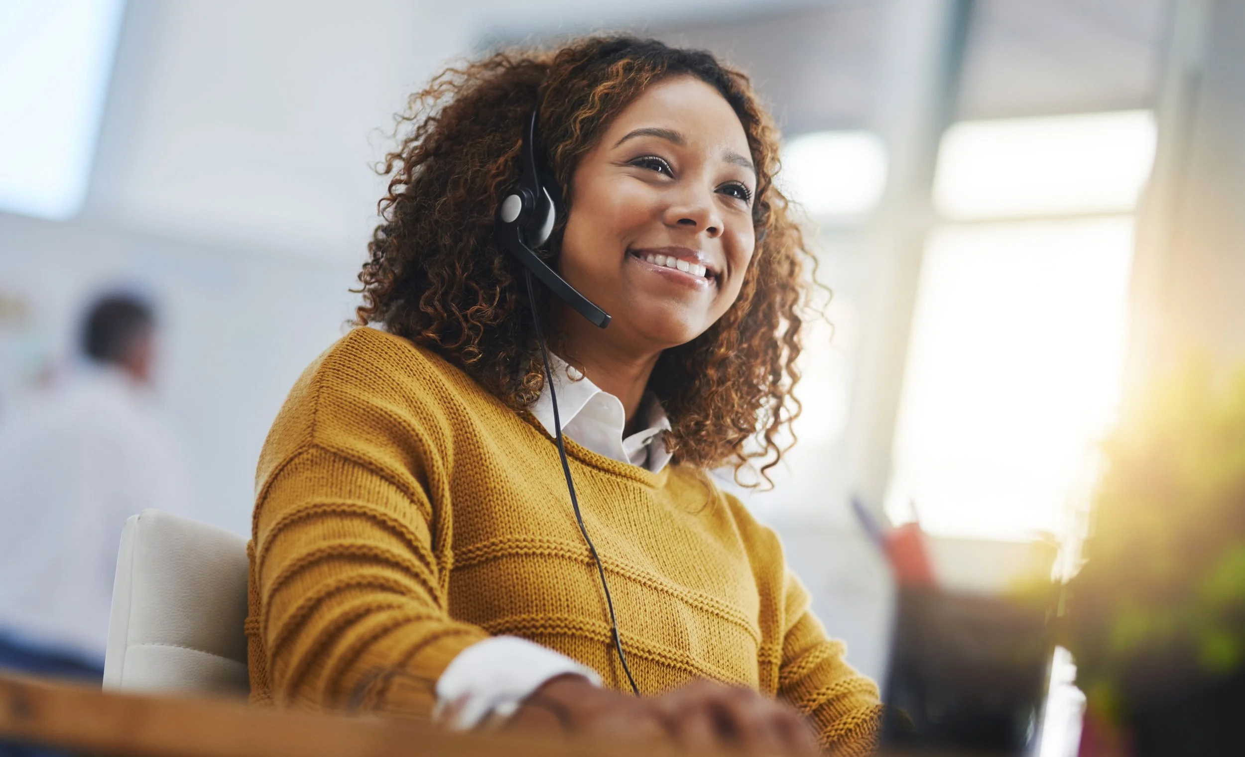 A woman with curly hair wearing a yellow sweater and a white collared shirt, smiling while working at a computer in an office environment.