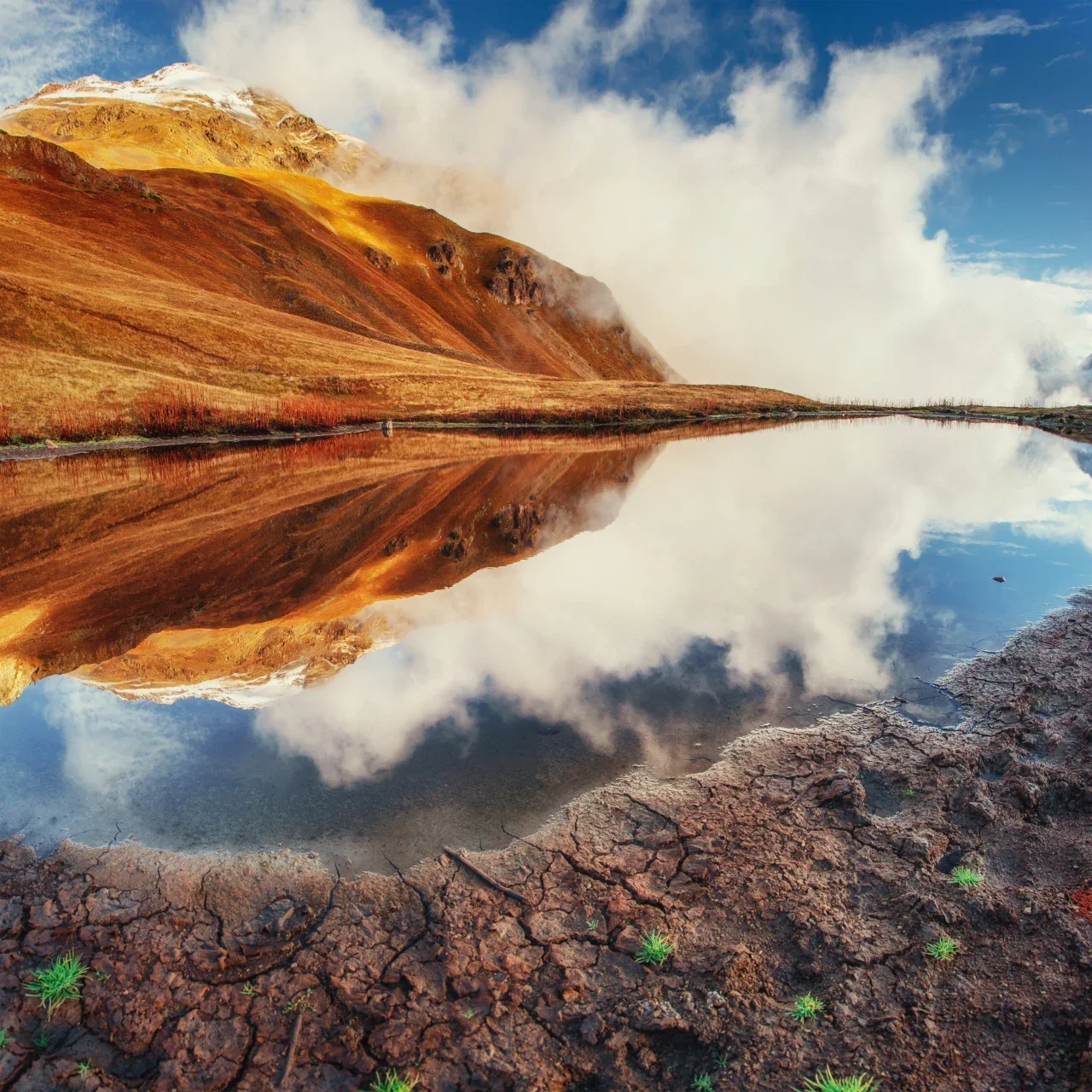 Scenic mountain landscape with a body of water reflecting the mountains and clouds, with cracked earth and small green plants in the foreground.
