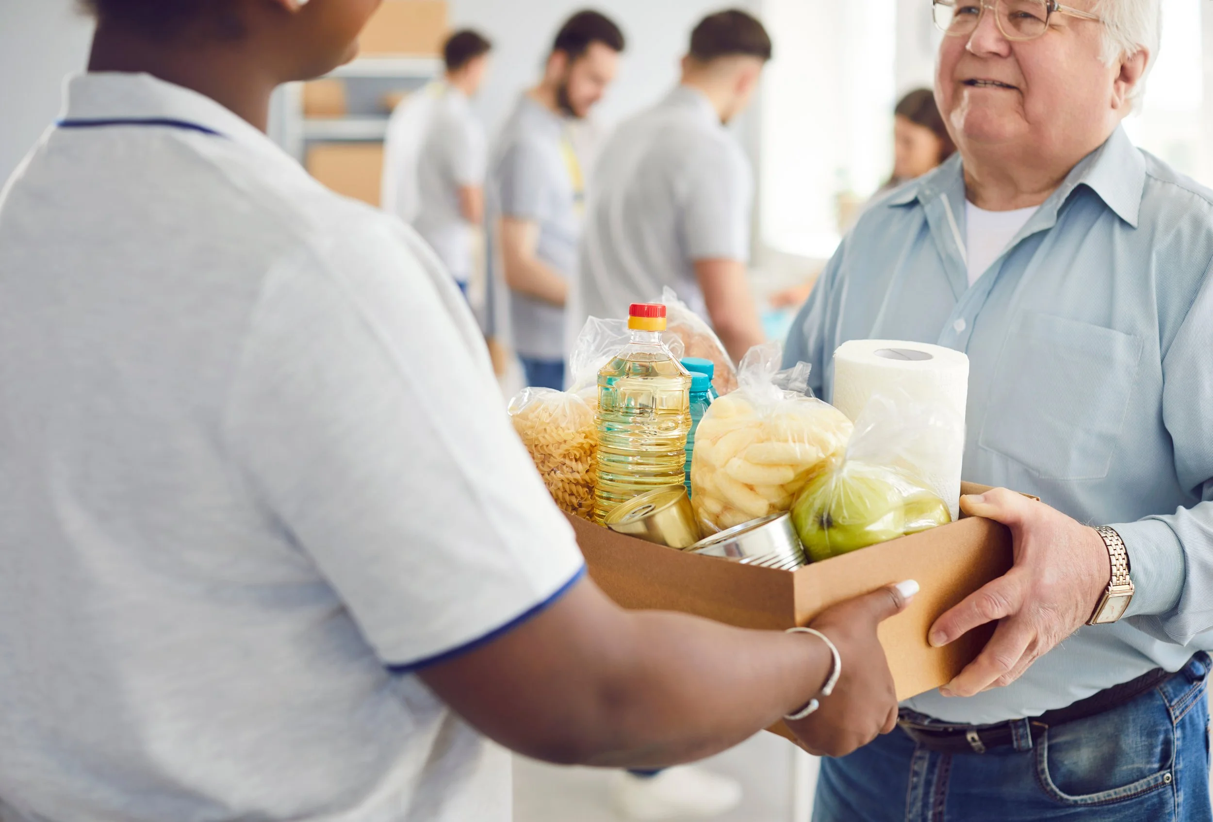 Two people exchanging a box of grocery items, including canned food, pasta, apples, and bottled drink, with several people in the background in what appears to be a community or food bank setting.
